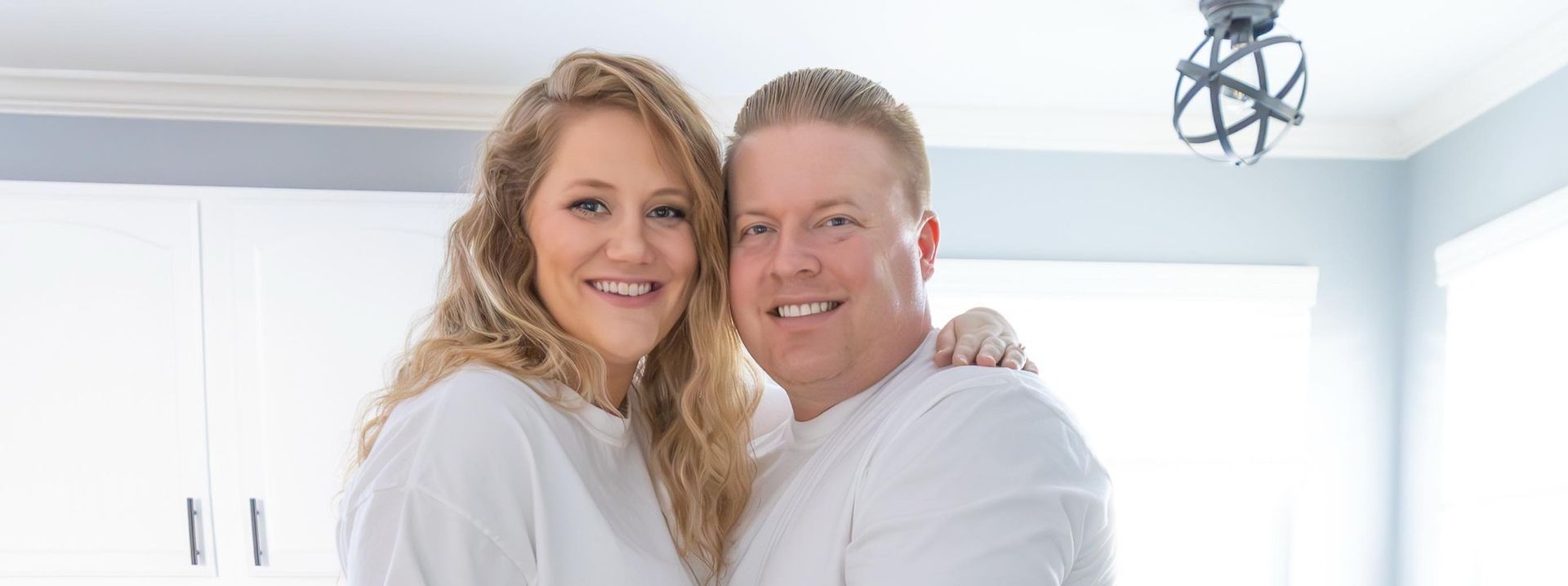 A smiling couple embraces indoors, likely in their home, wearing white shirts.
