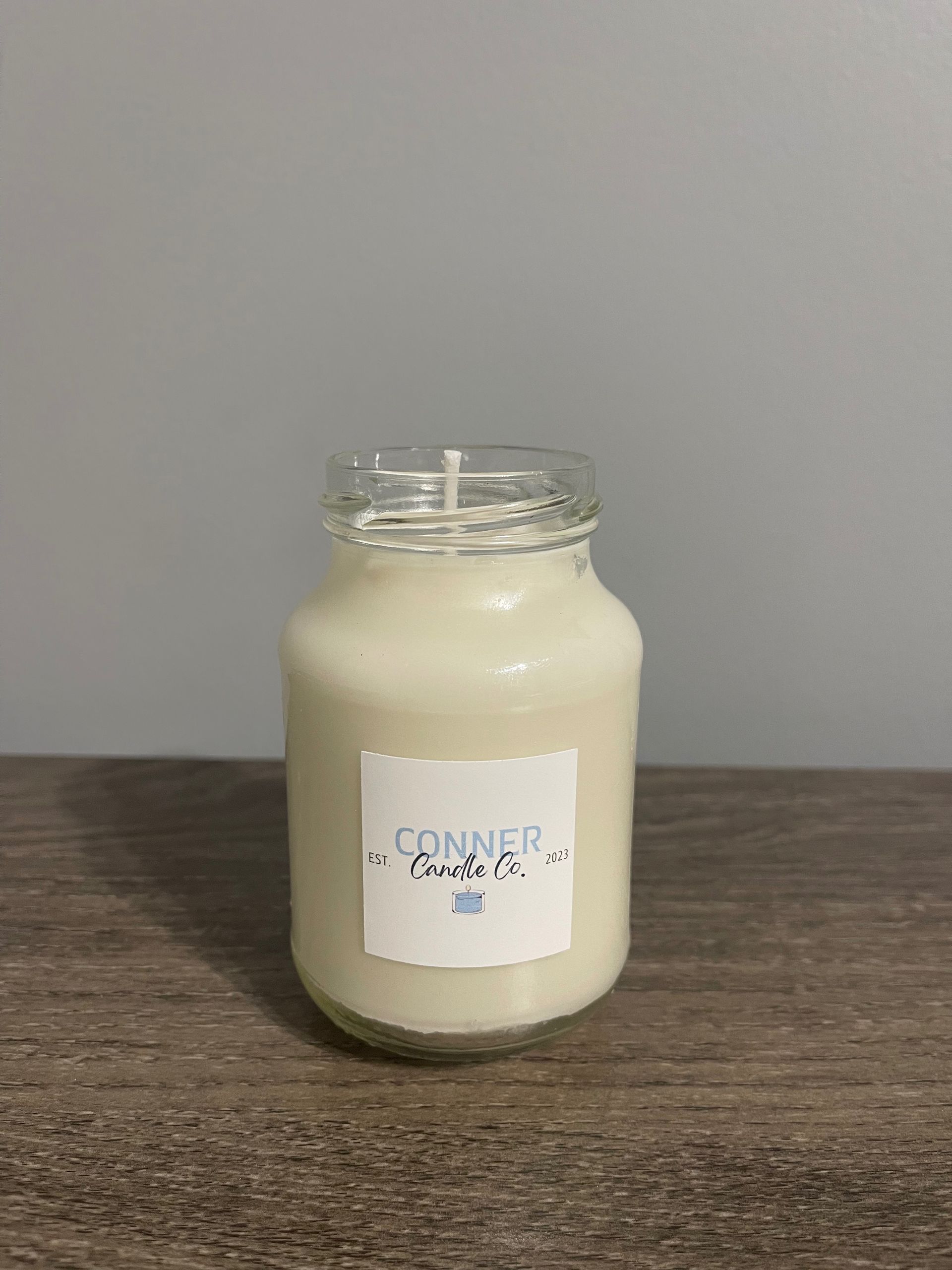 White candle in a jar with a label, sitting on a wooden surface against a grey background.