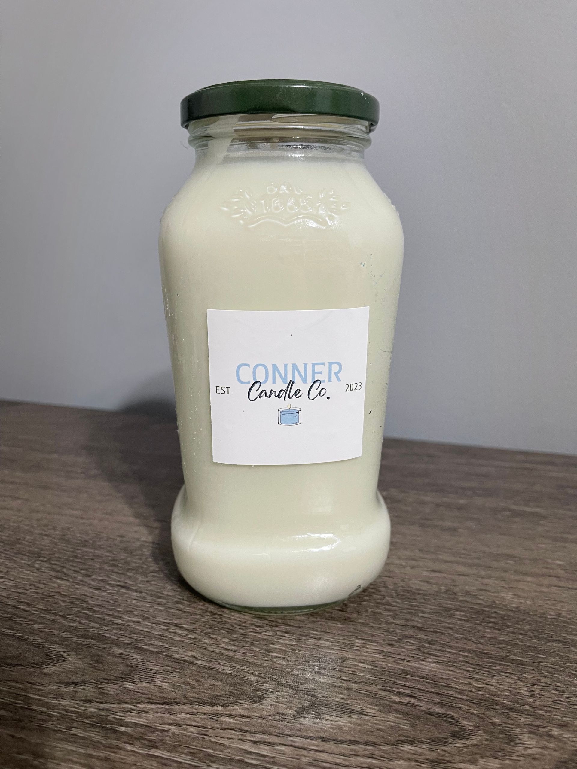 White candle in a glass jar with a green lid; label reads 