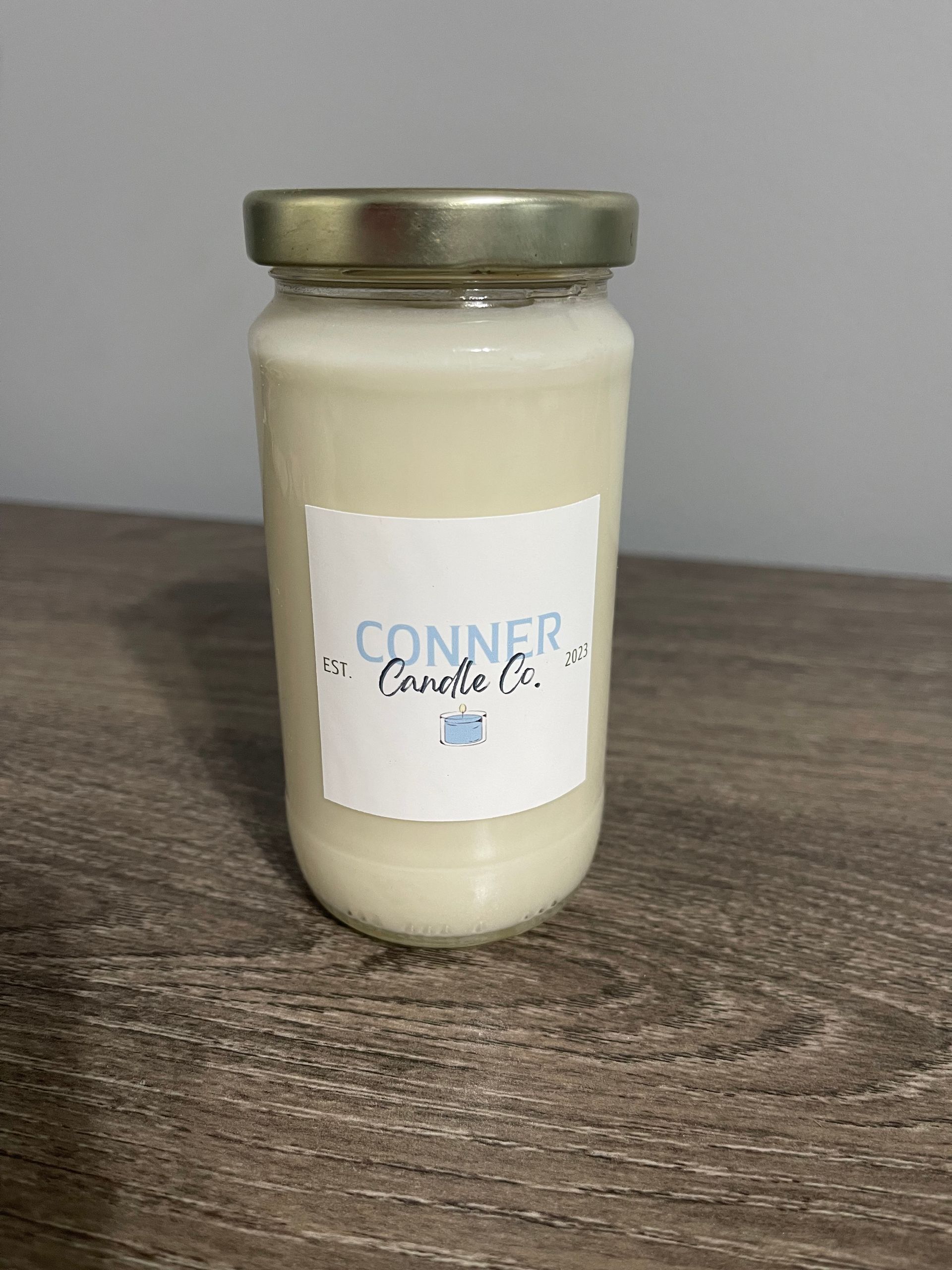 A white candle in a glass jar with a gold lid on a wooden surface. Label reads 