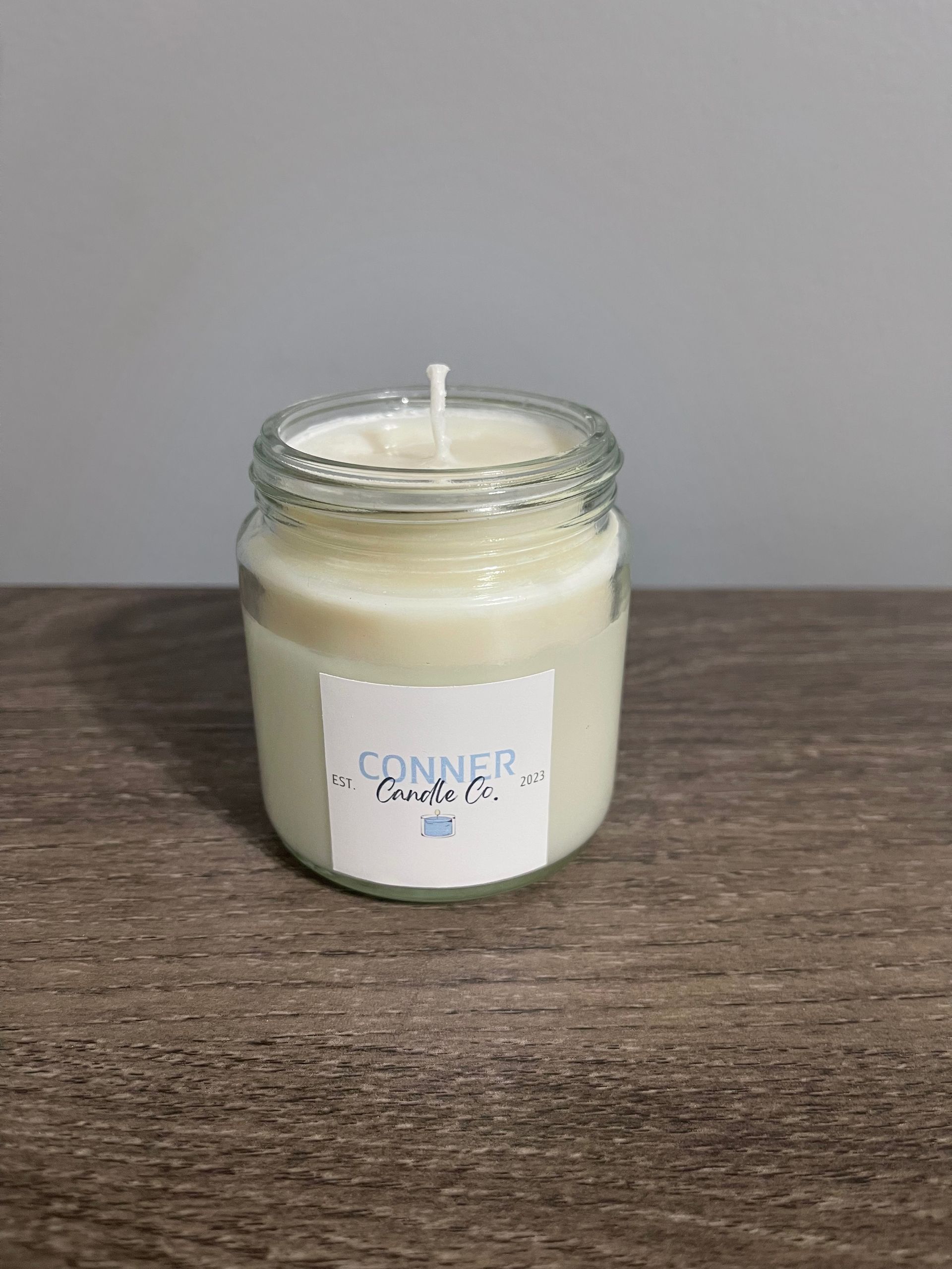 White candle in a clear glass jar with a label, sitting on a wooden surface.