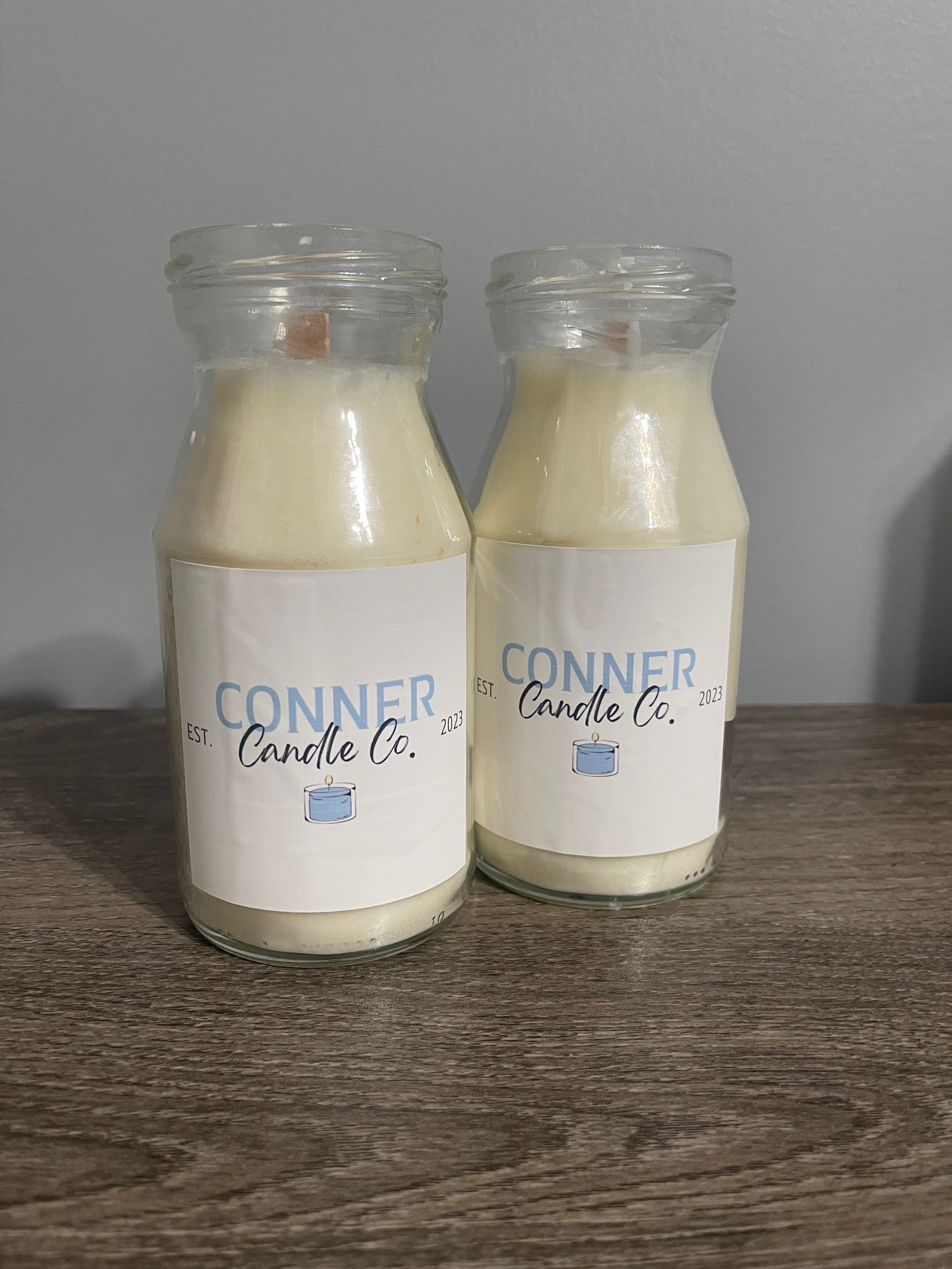 Two cream-colored candles in glass milk bottle jars with blue and white labels; the label reads, 