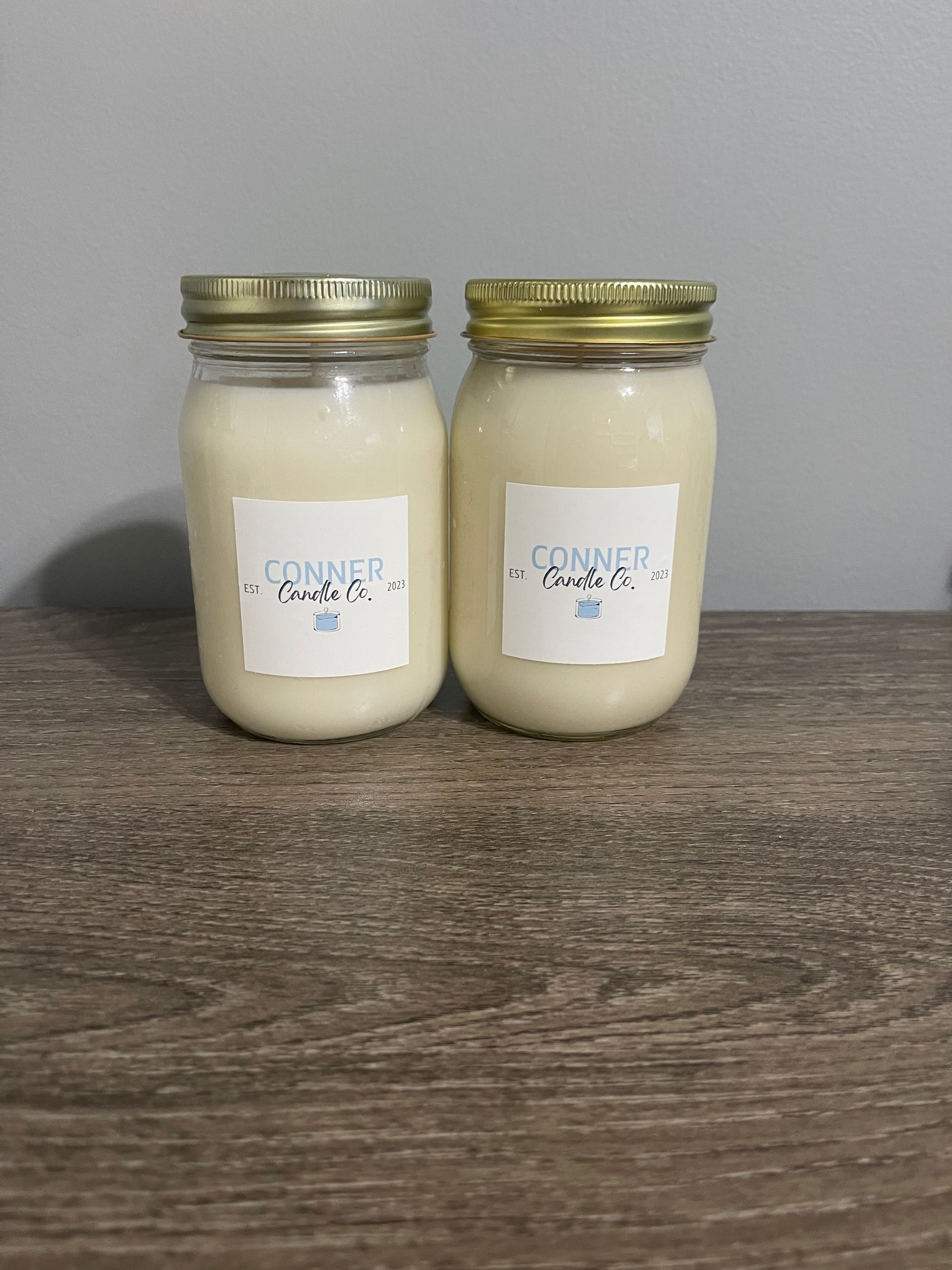 Two mason jars filled with off-white wax, gold lids, and white labels on a wooden surface.