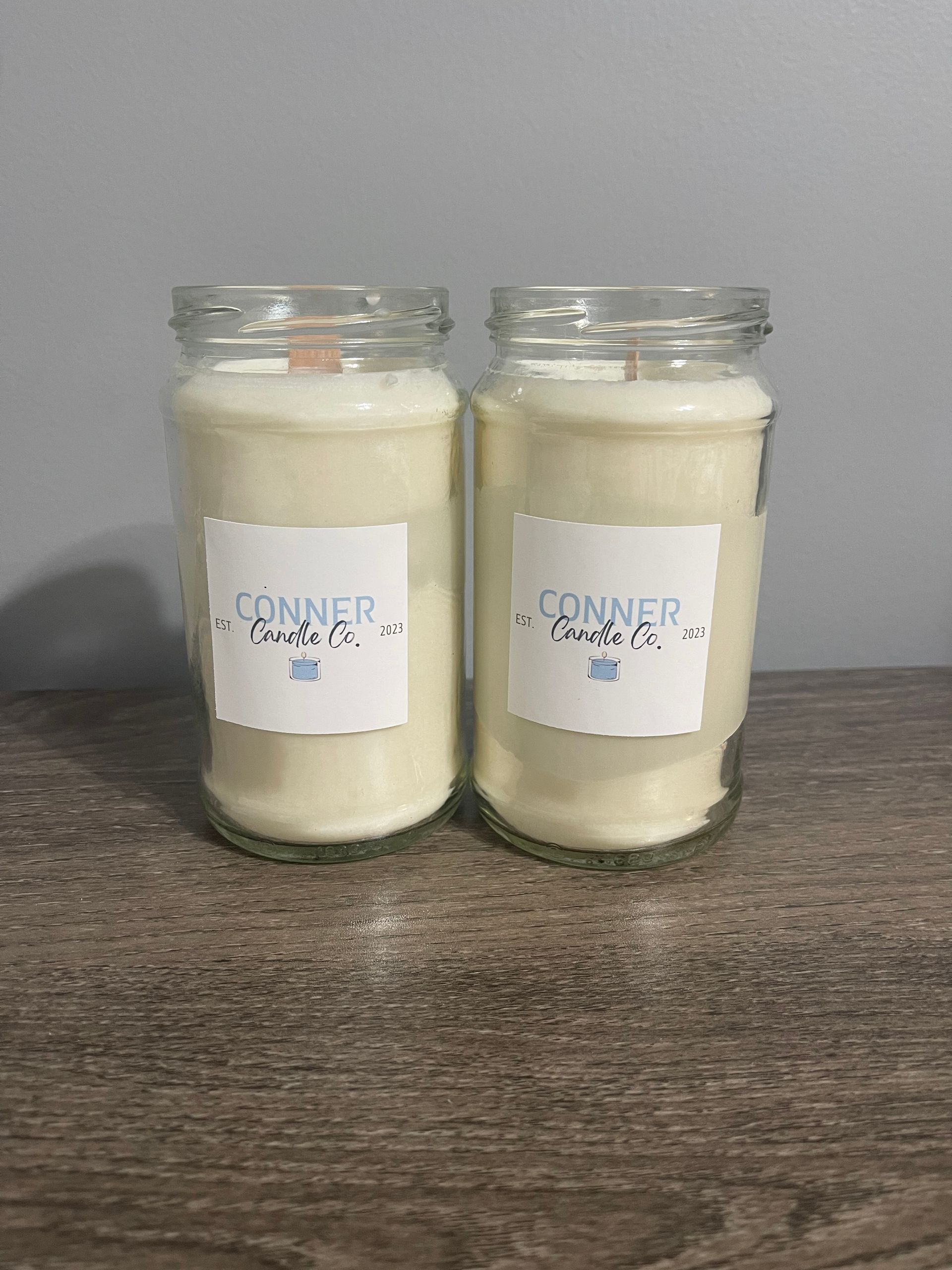 Two cream-colored candles in glass jars with white labels, on a wooden surface.