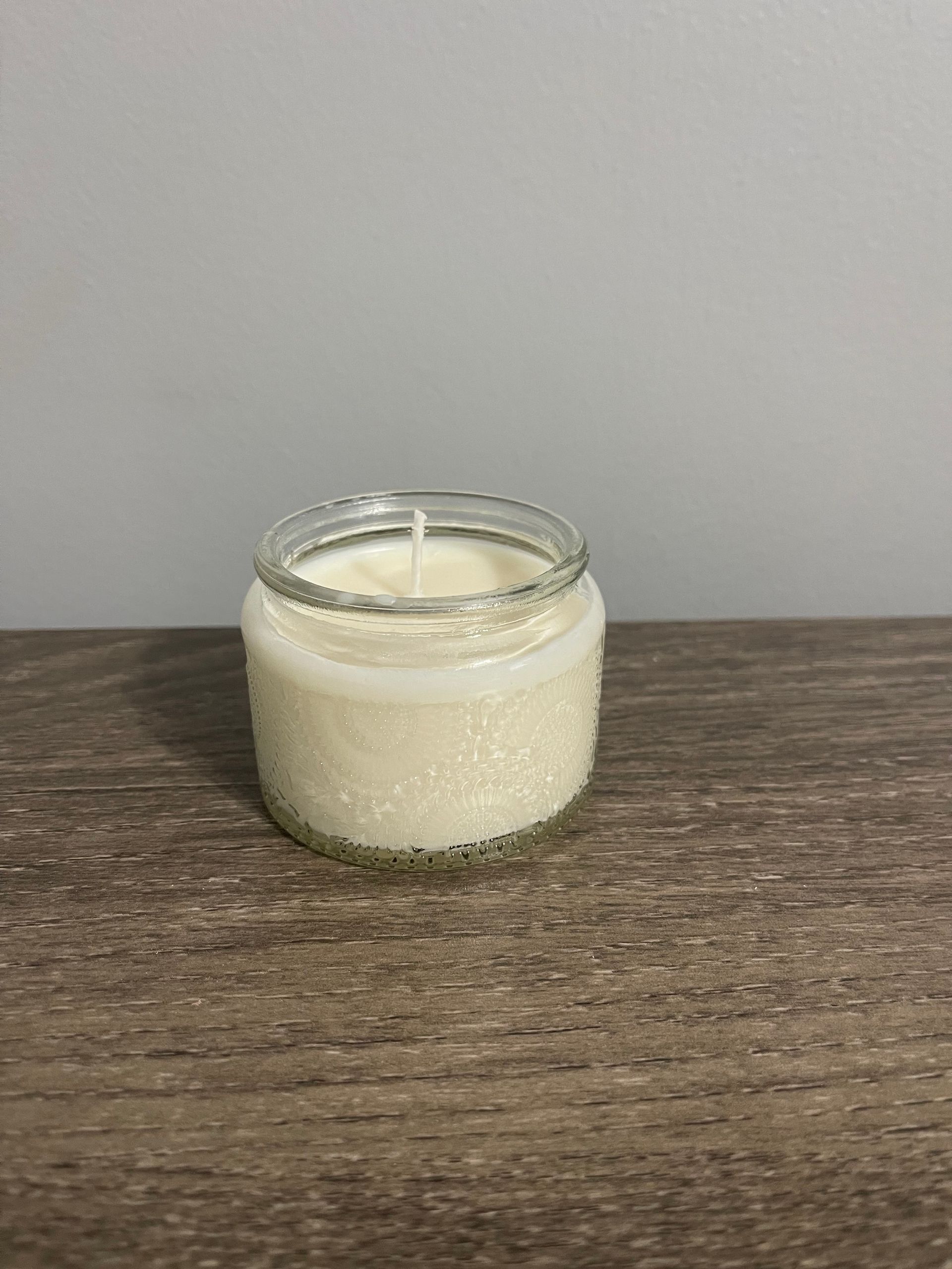 White candle in a clear glass jar on a wooden surface, against a grey background.
