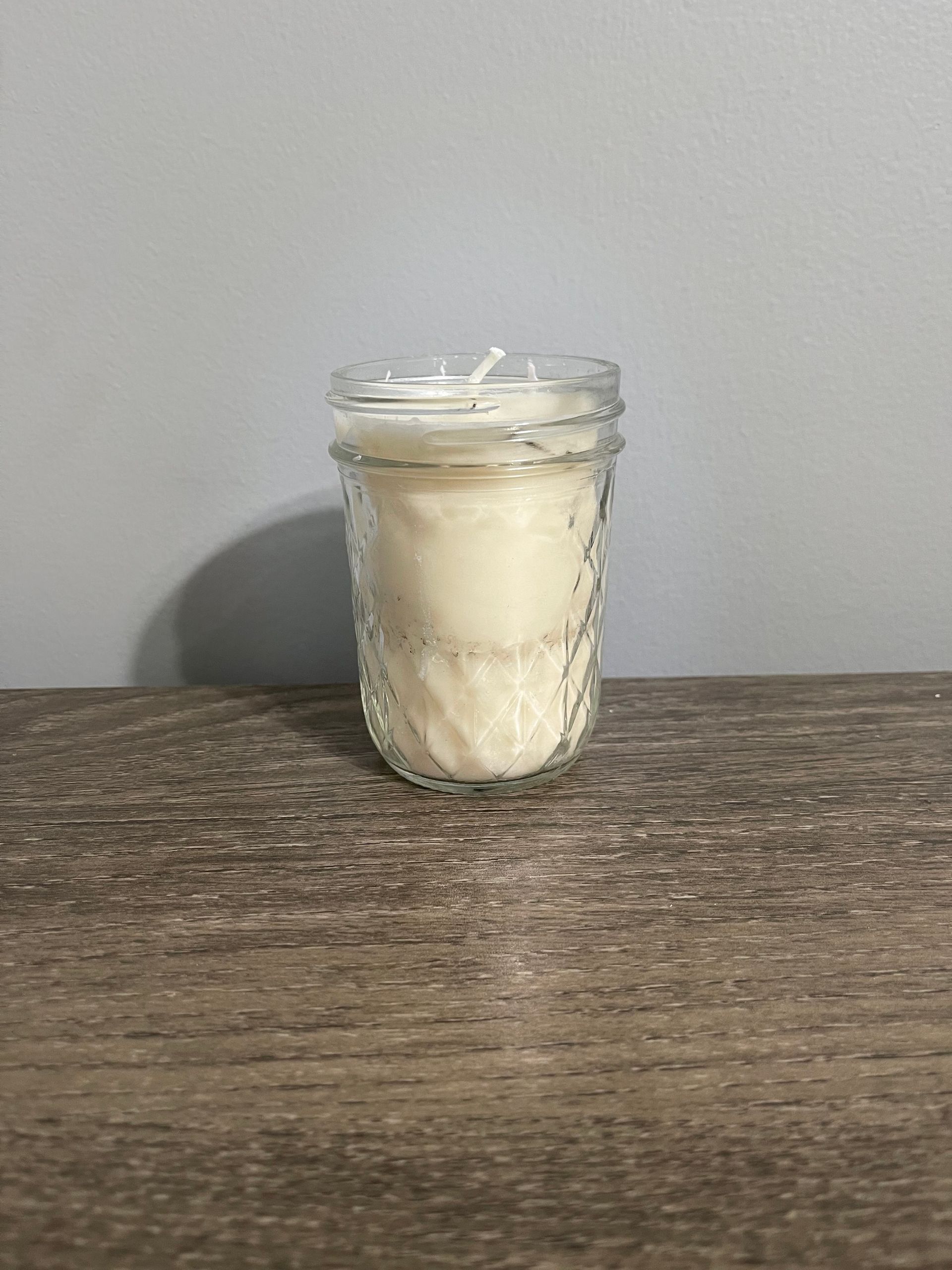 Cream-colored candle in a textured glass jar on a wooden surface, with a pale blue wall background.