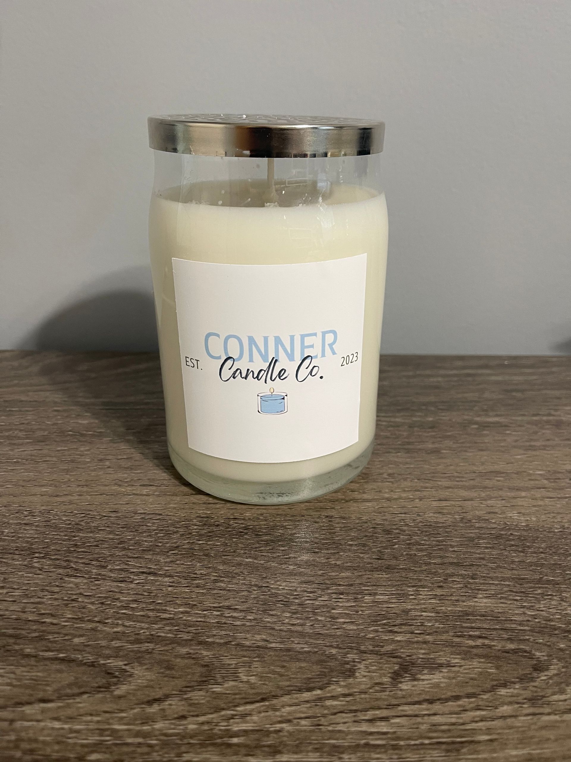 A candle in a clear glass jar with a silver lid sits on a wooden surface. Label reads 