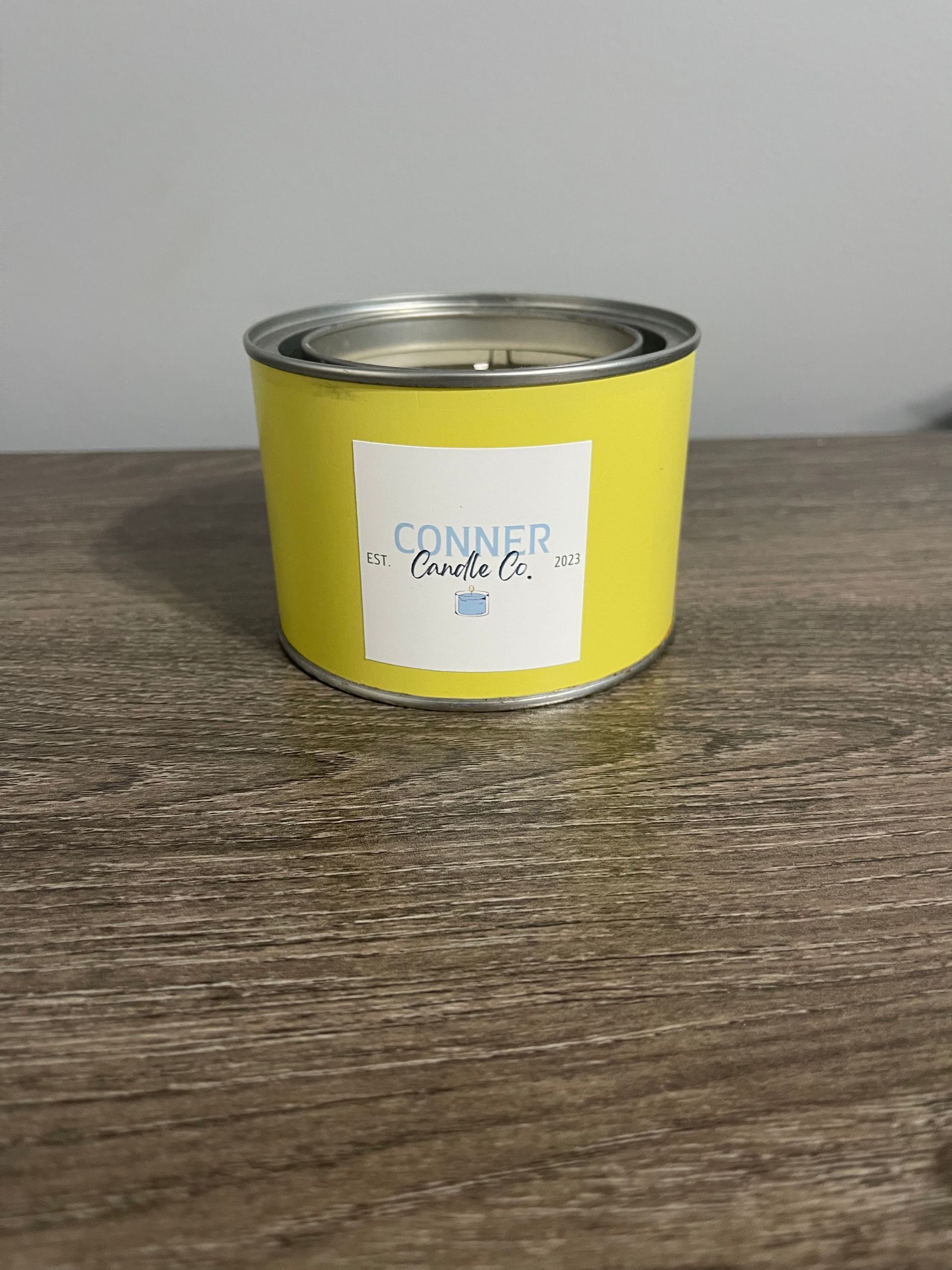 Yellow candle in a metal container on a wooden surface. The label says “Conner Candle Co.”