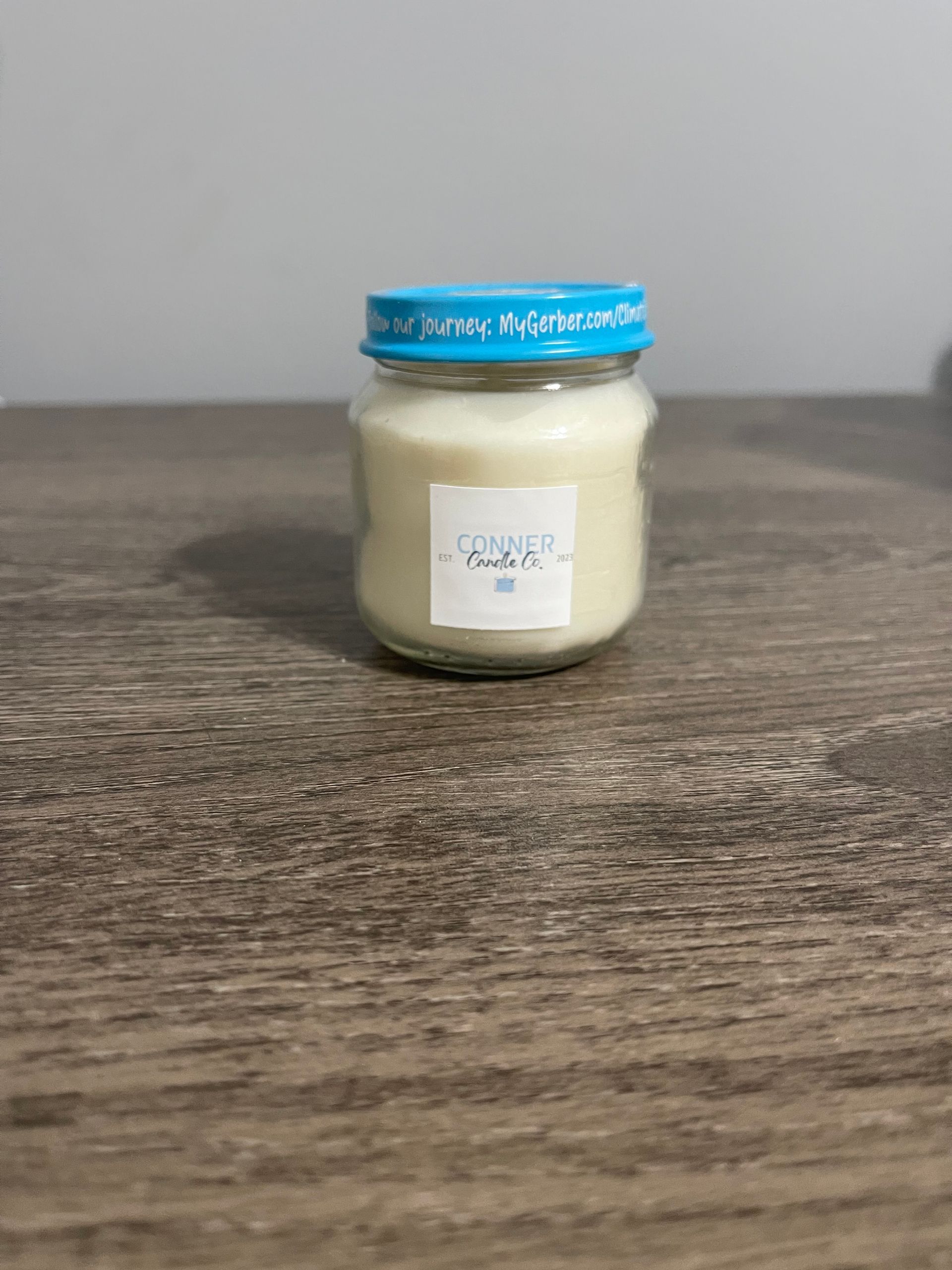 A small candle in a baby food jar with a blue lid, sitting on a wooden table.