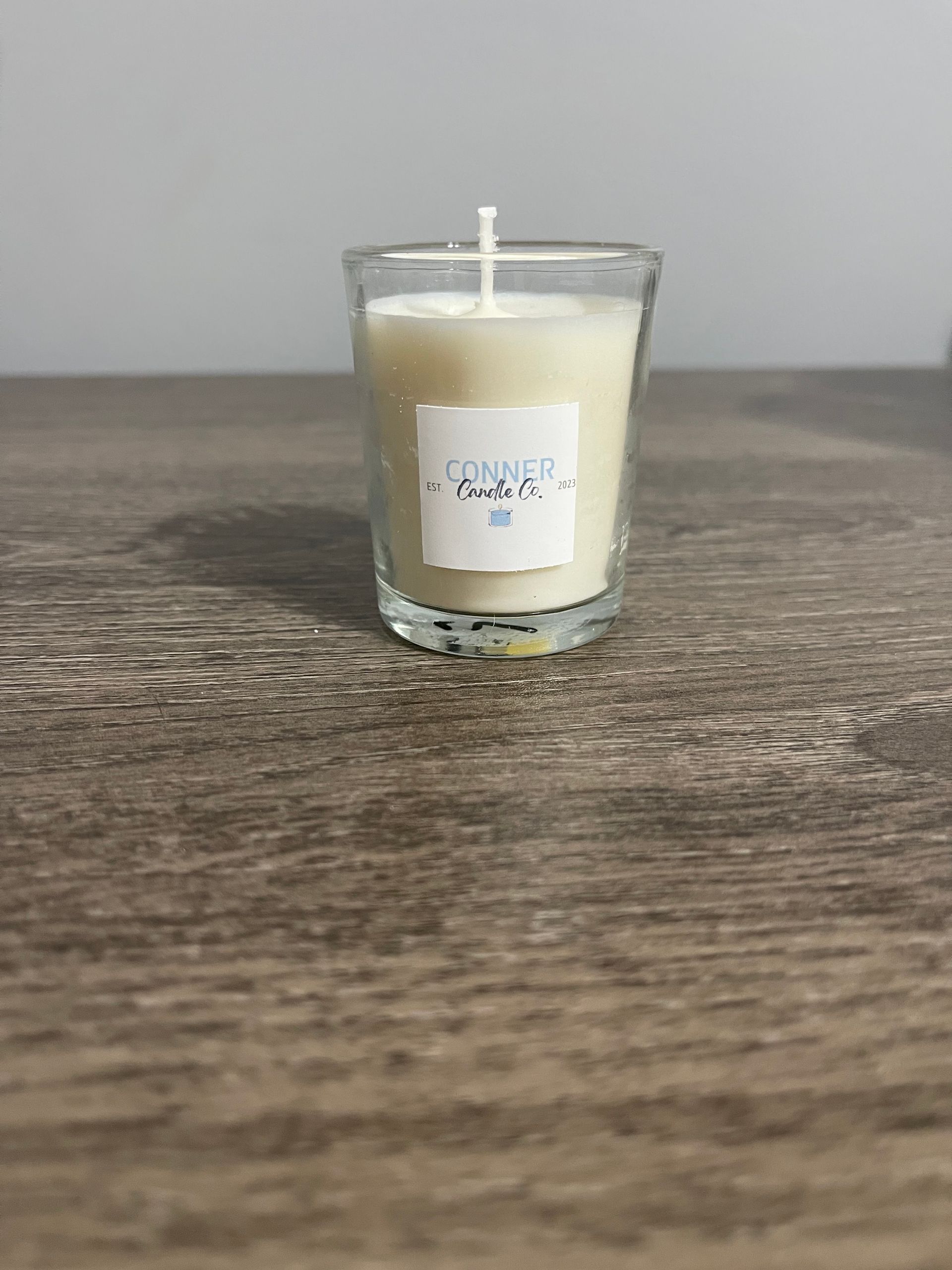 A lit white candle in a clear glass on a wooden surface, with a small label.
