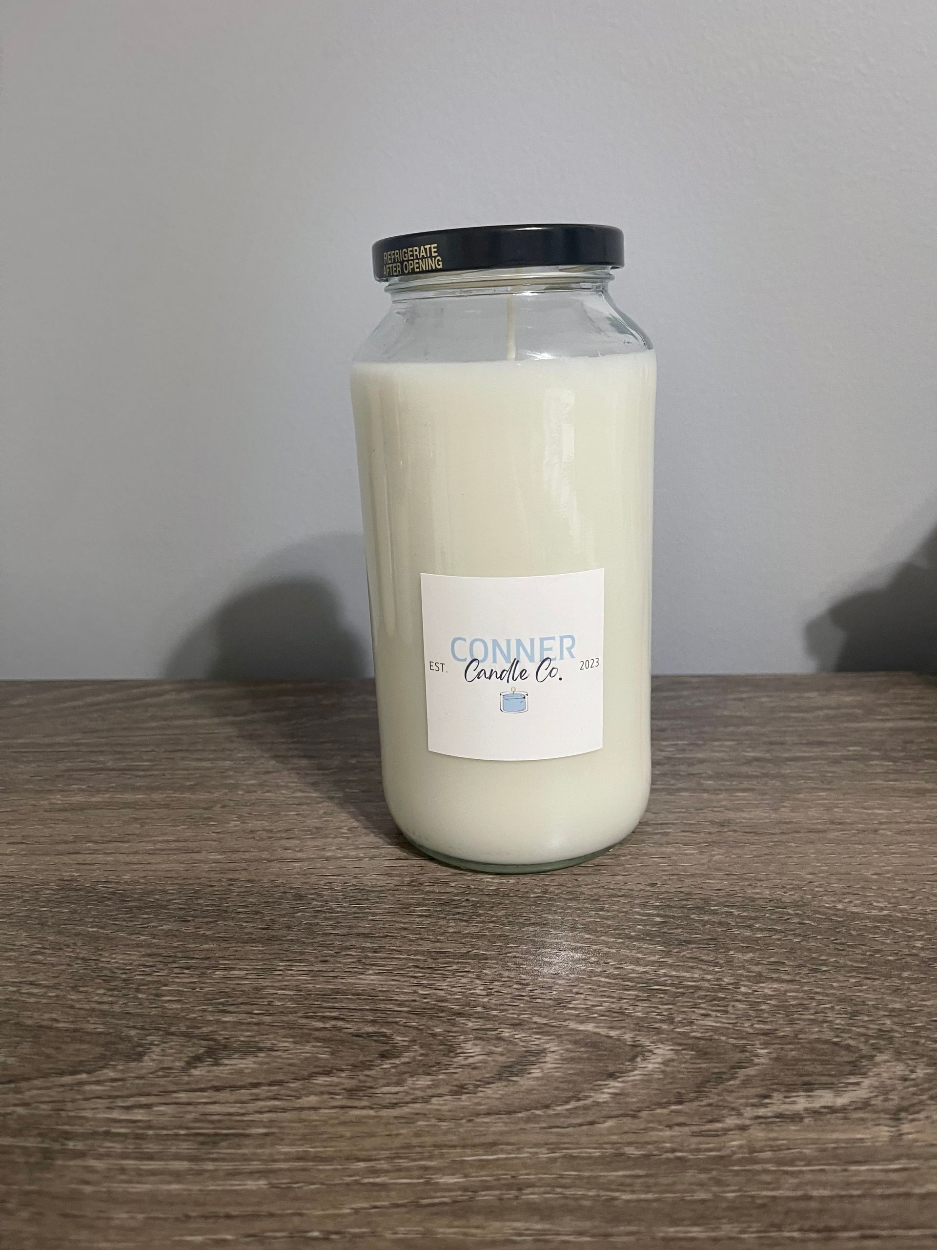 A white candle in a clear glass jar with a black lid sits on a wooden surface.