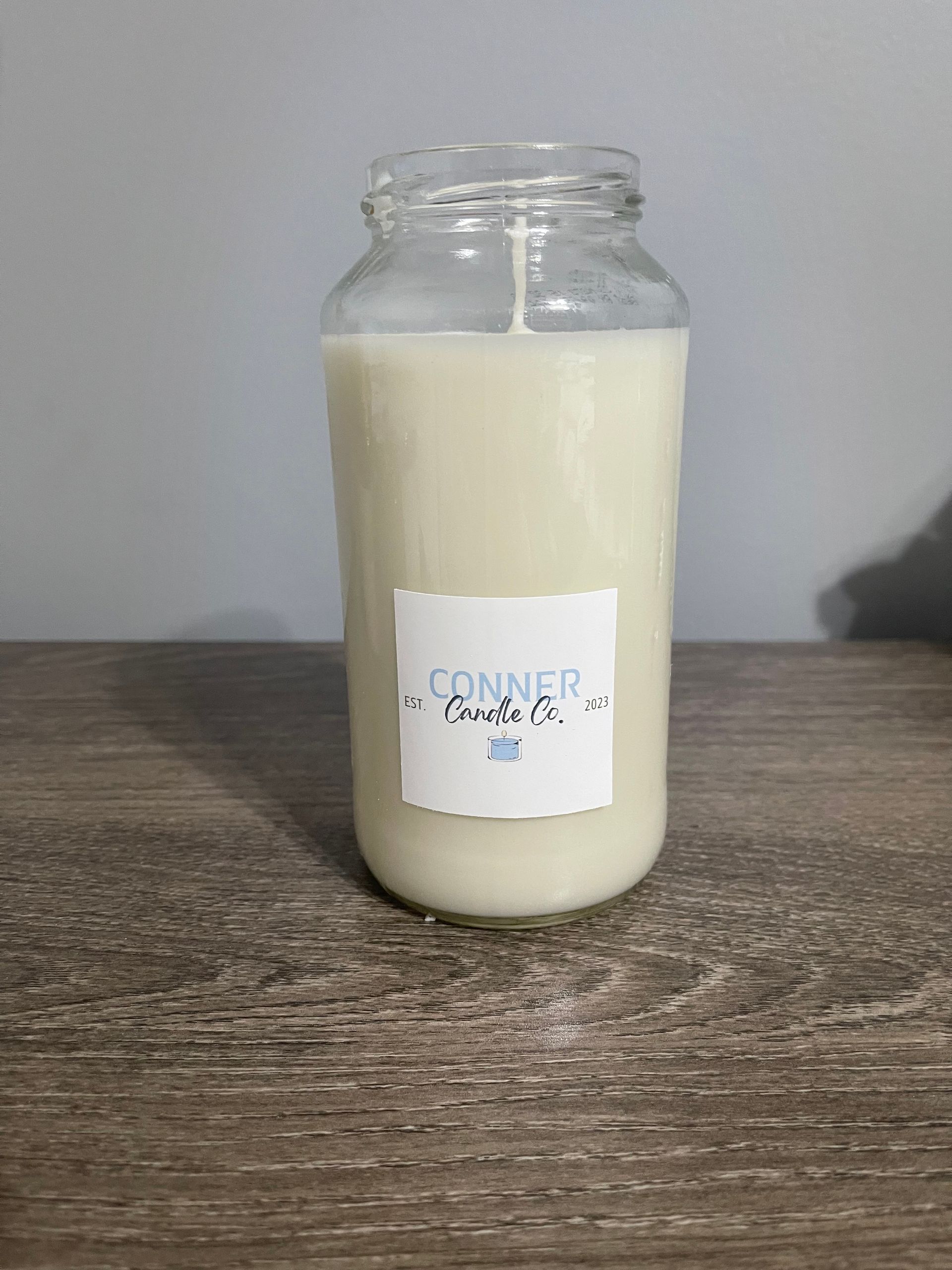 Candle in a clear glass jar with a label, sitting on a wooden surface against a gray wall.