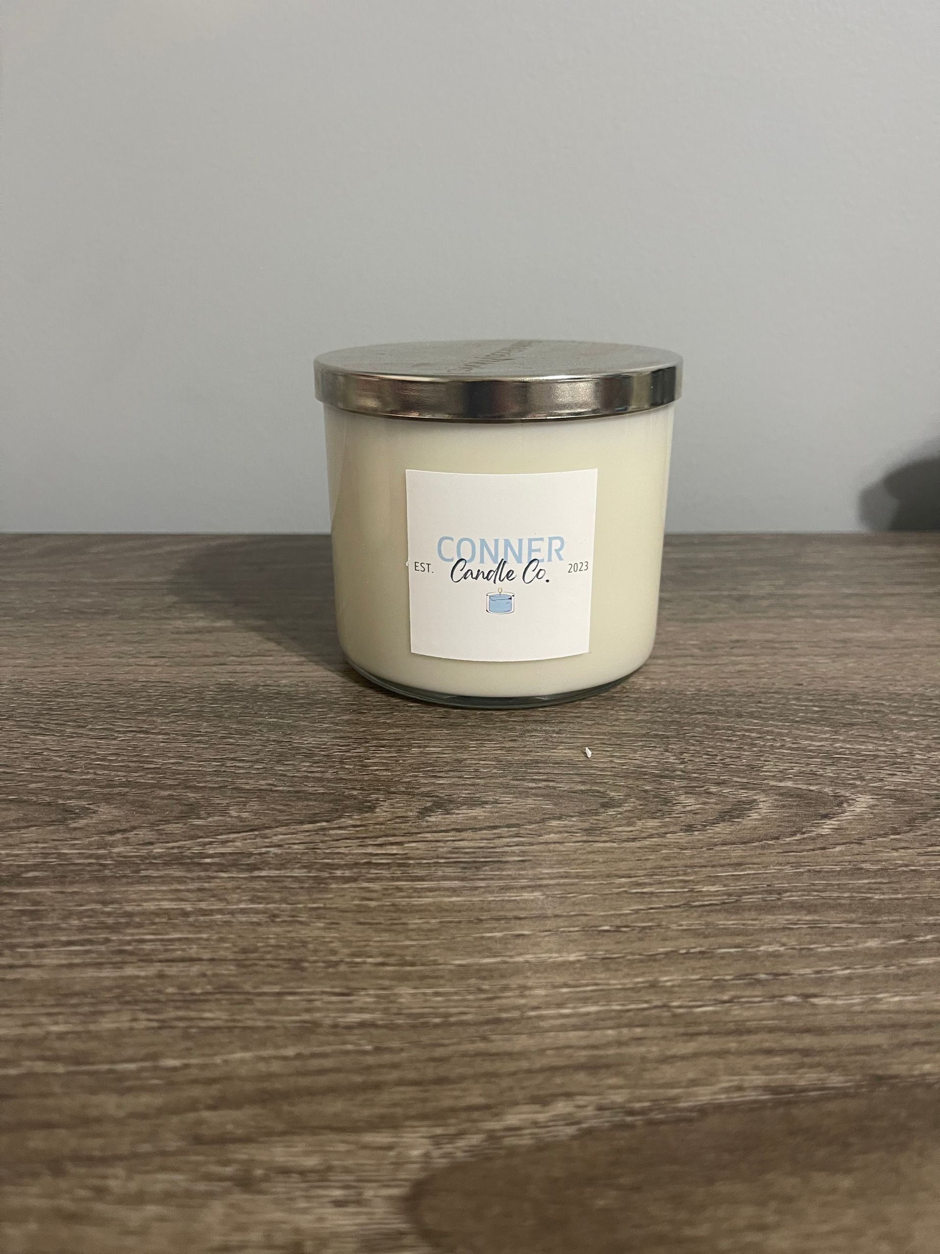 White candle in glass jar with silver lid, sitting on a wood surface, with a label that says 