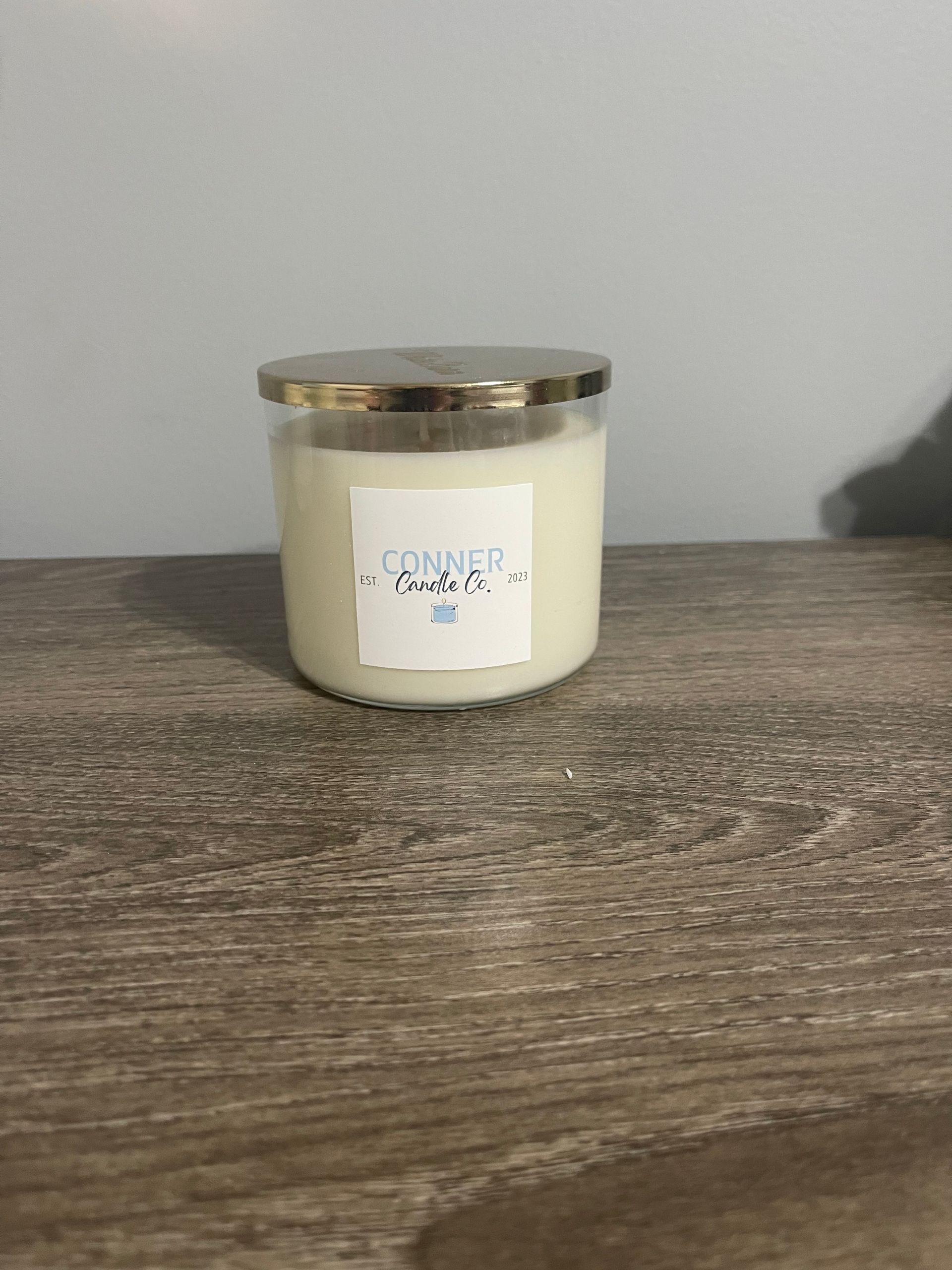 Candle in a glass jar with gold lid, sits on a wooden surface. A small white label is on the jar.