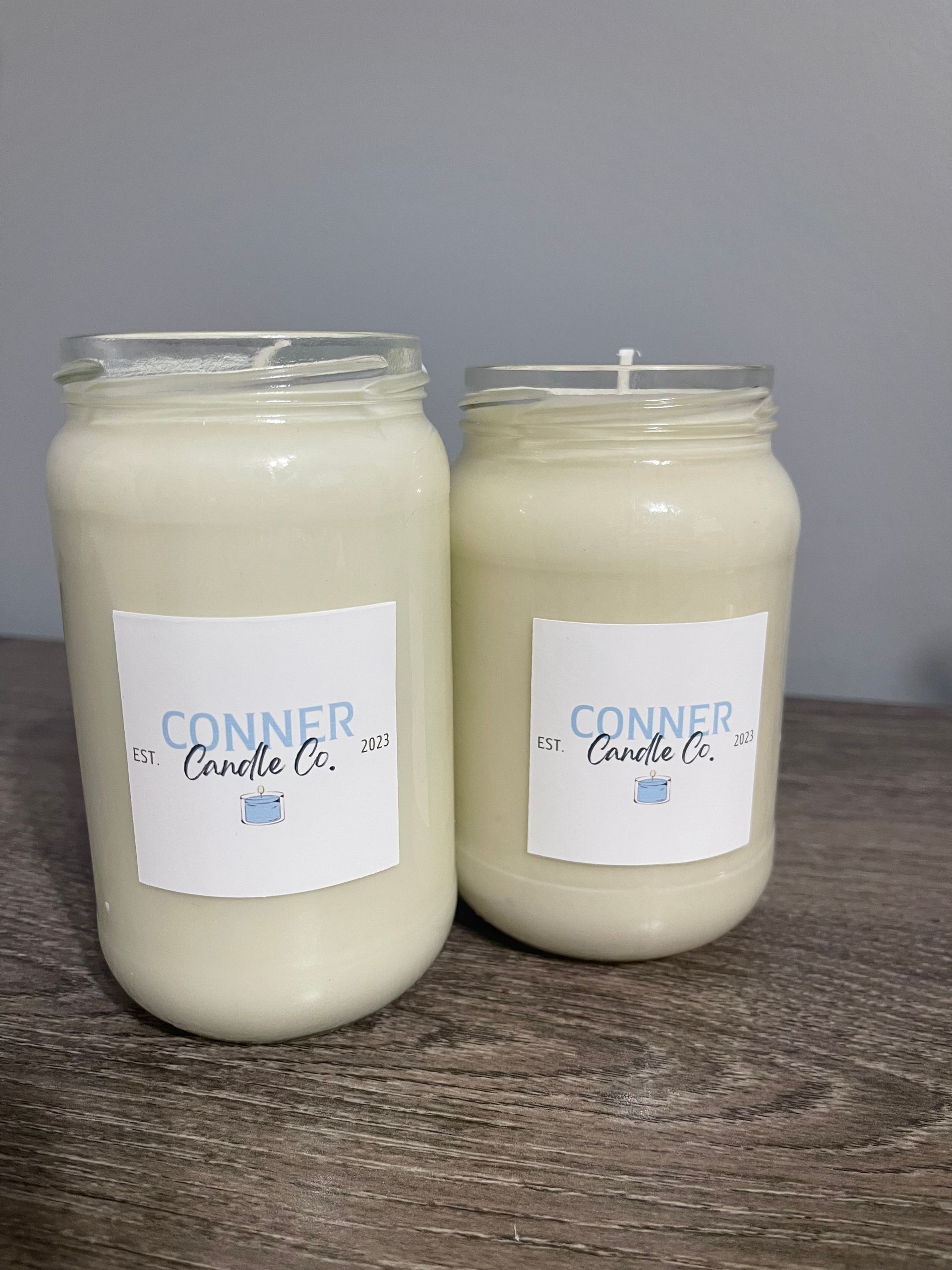 Two cream-colored jar candles with white labels. One candle is taller than the other, set on a wooden surface.