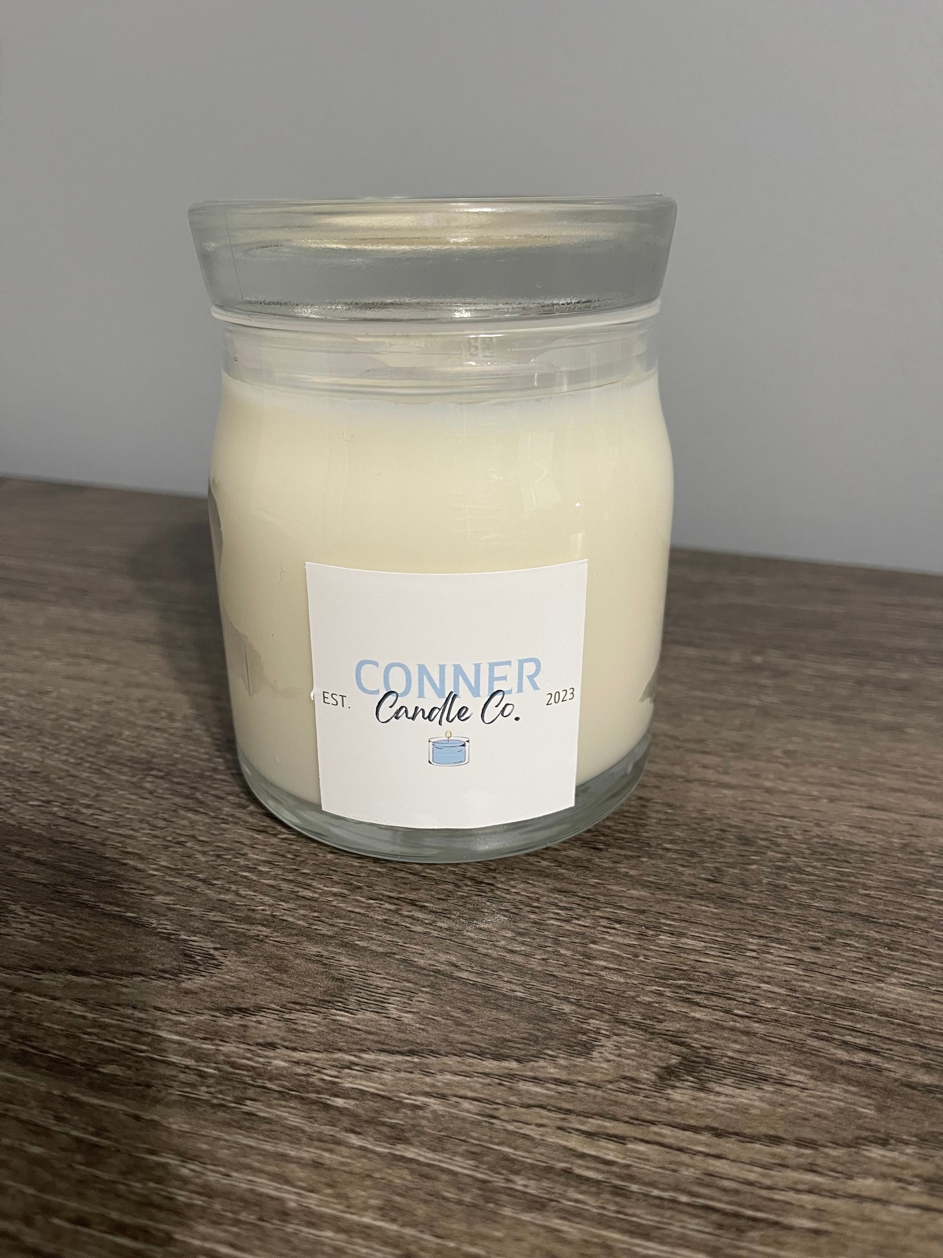 White candle in a clear glass jar with a white label that has black text, resting on a brown wooden surface.
