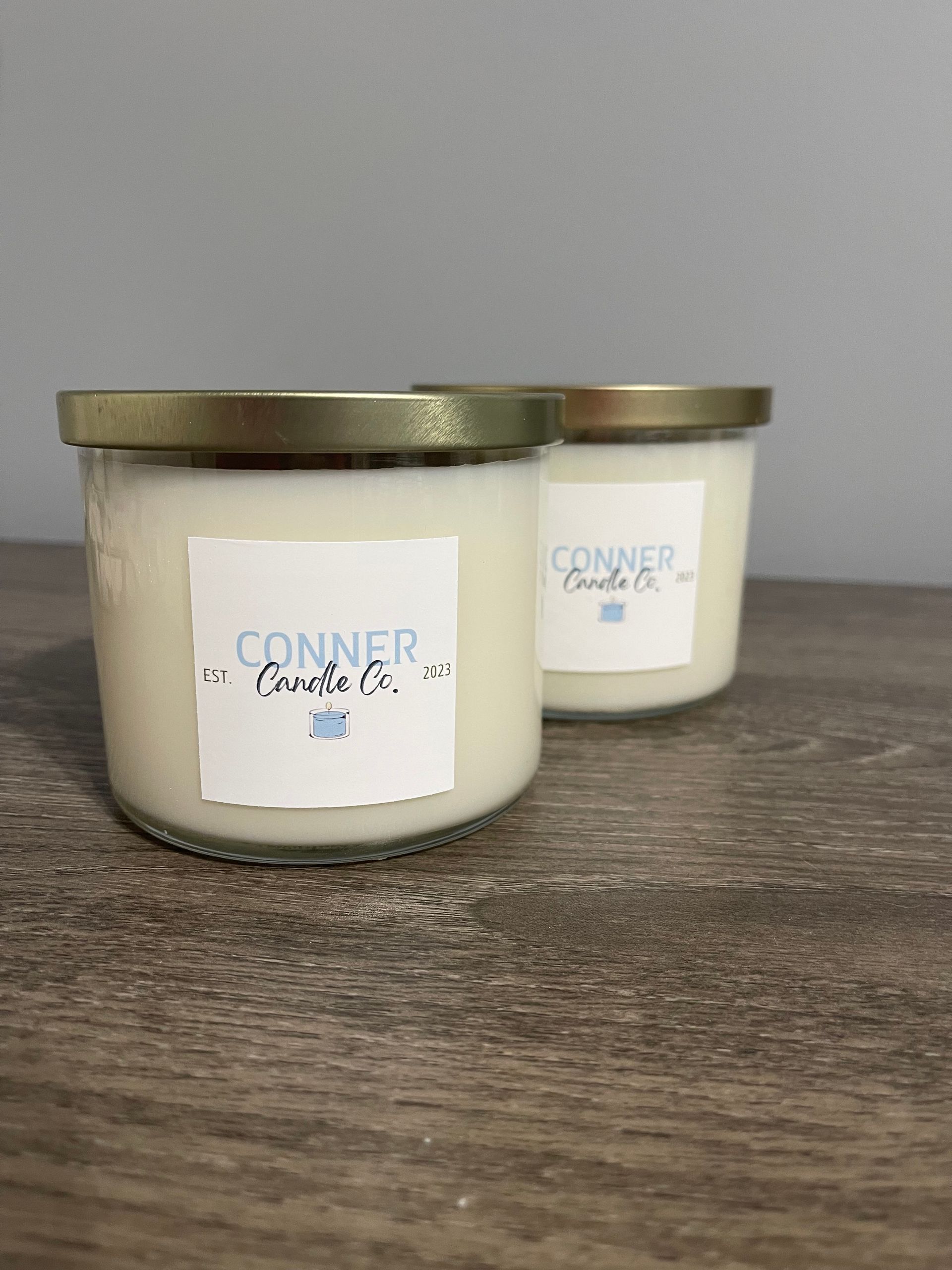 Two Conner Candle Co. candles with gold lids and white labels on a wooden surface.