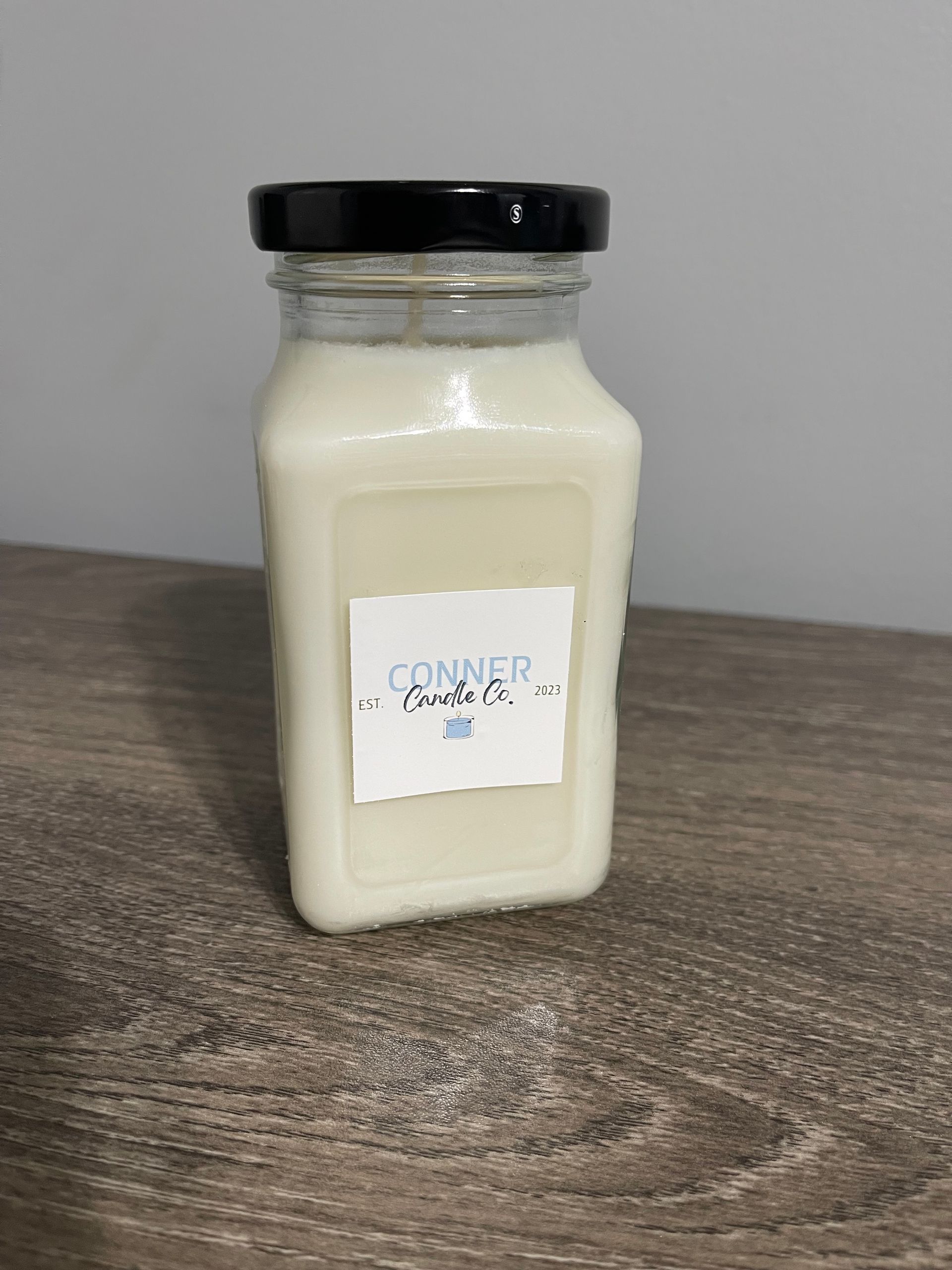 White candle in a square glass jar with a black lid; a label reads 