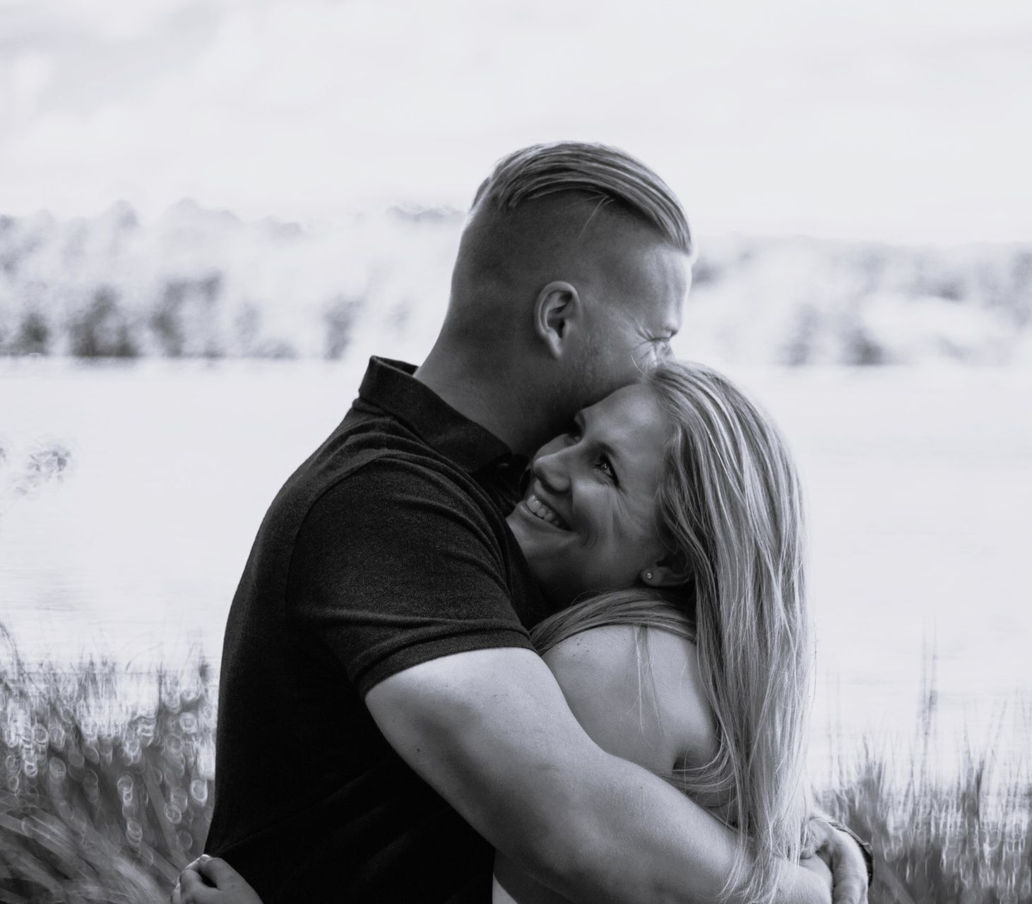 Man hugs woman, kissing her forehead. They stand near water, smiling. Black and white.