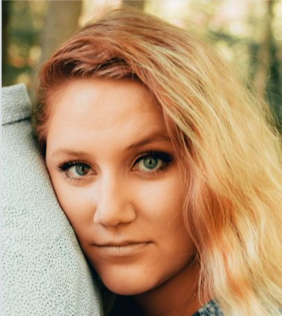 Woman with blonde hair, leaning head on shoulder; blue eyes, looking towards camera.