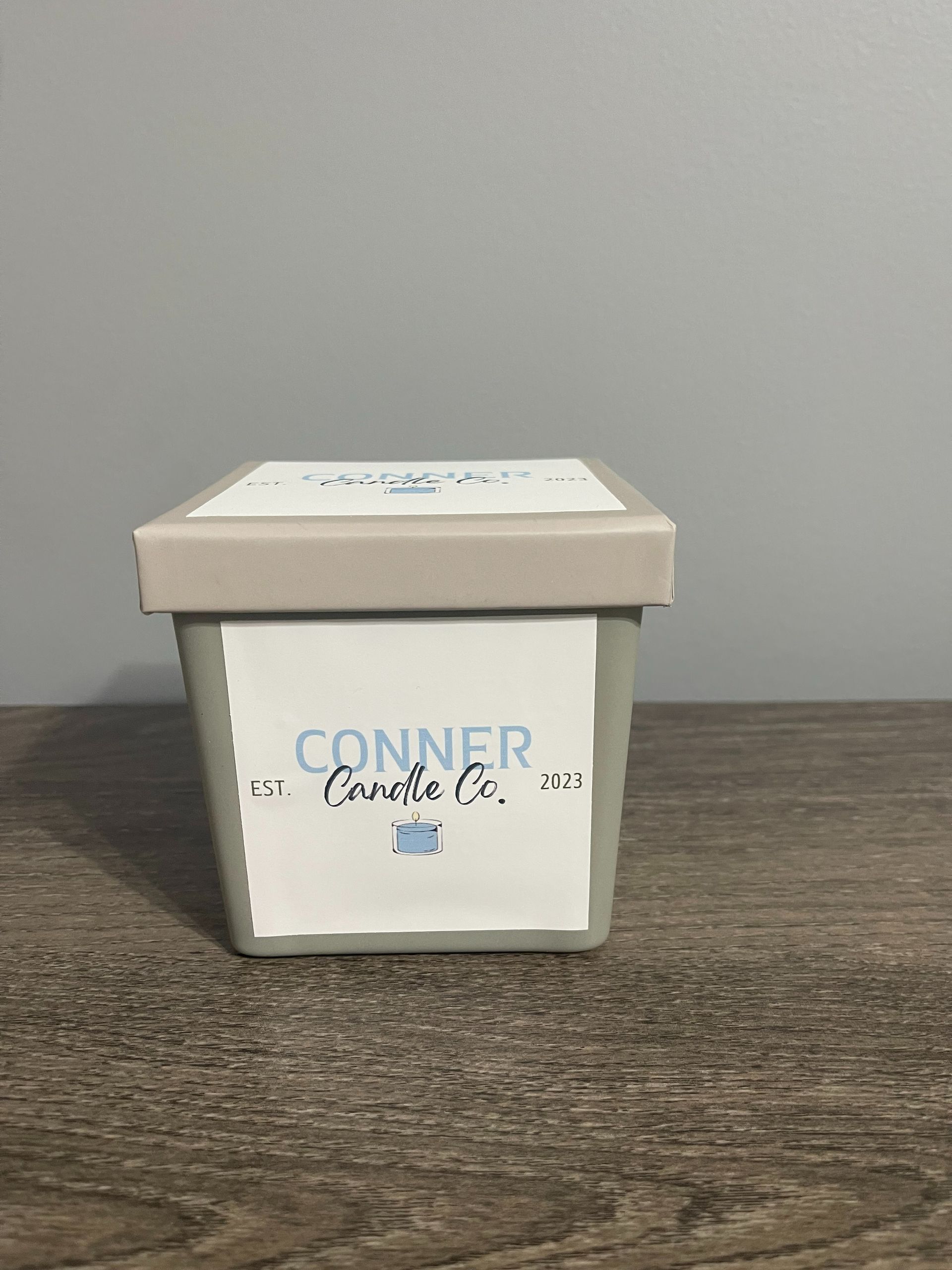 Gray square candle with a Conner Candles Co. label, set on a wooden surface against a gray wall.