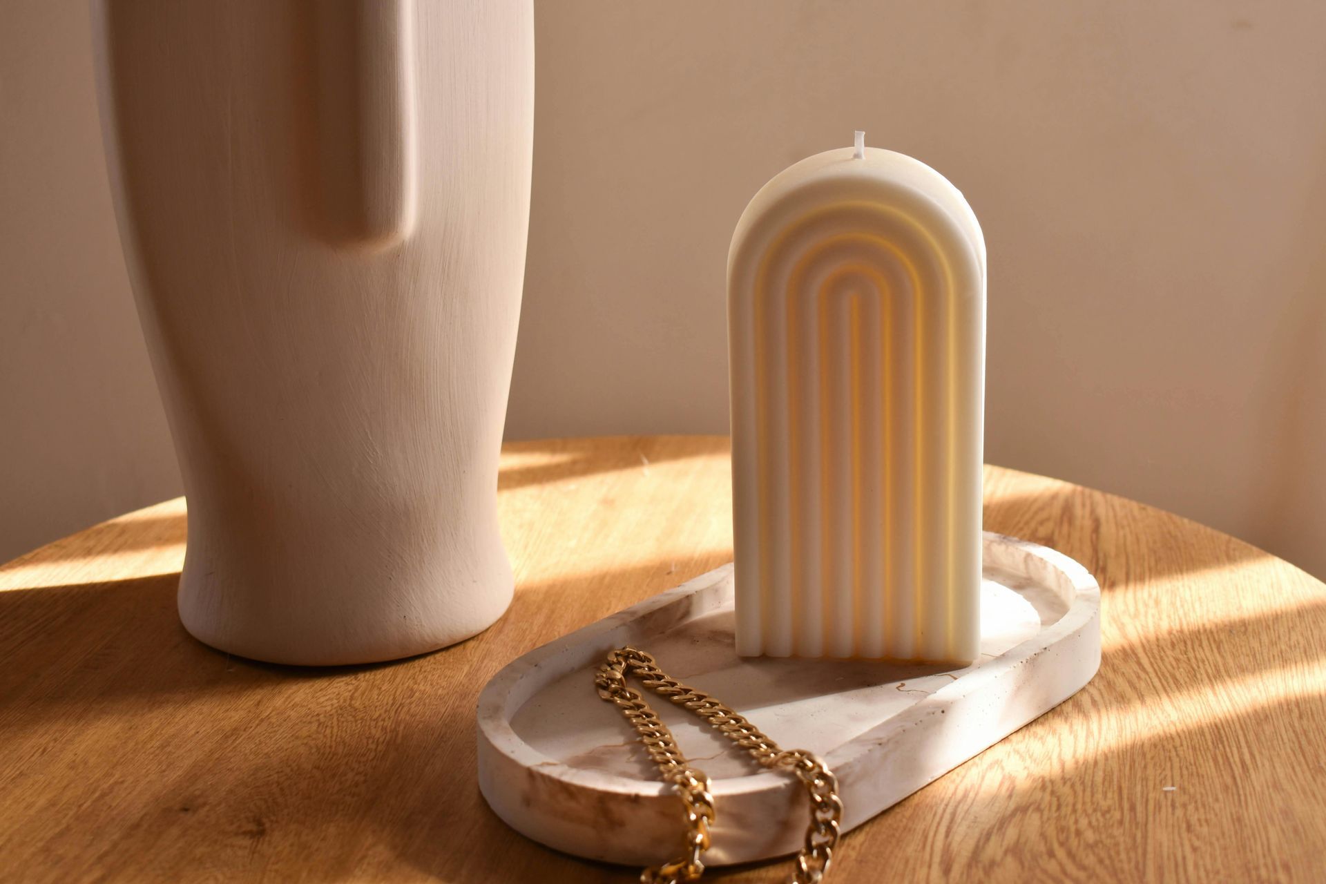 Off-white arch-shaped candle on a tray, next to a vase and gold necklace, on a wooden table.