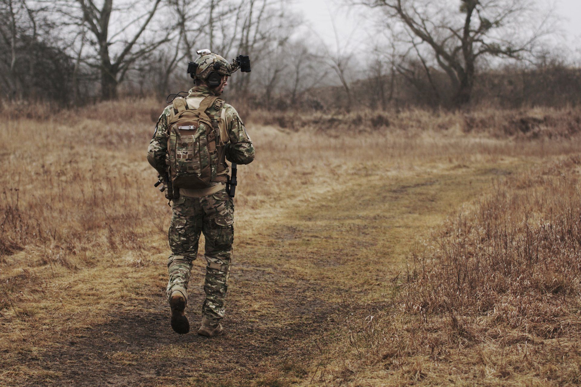 Soldier in camouflage walks on a dirt path through a field of brown grass, carrying a backpack.