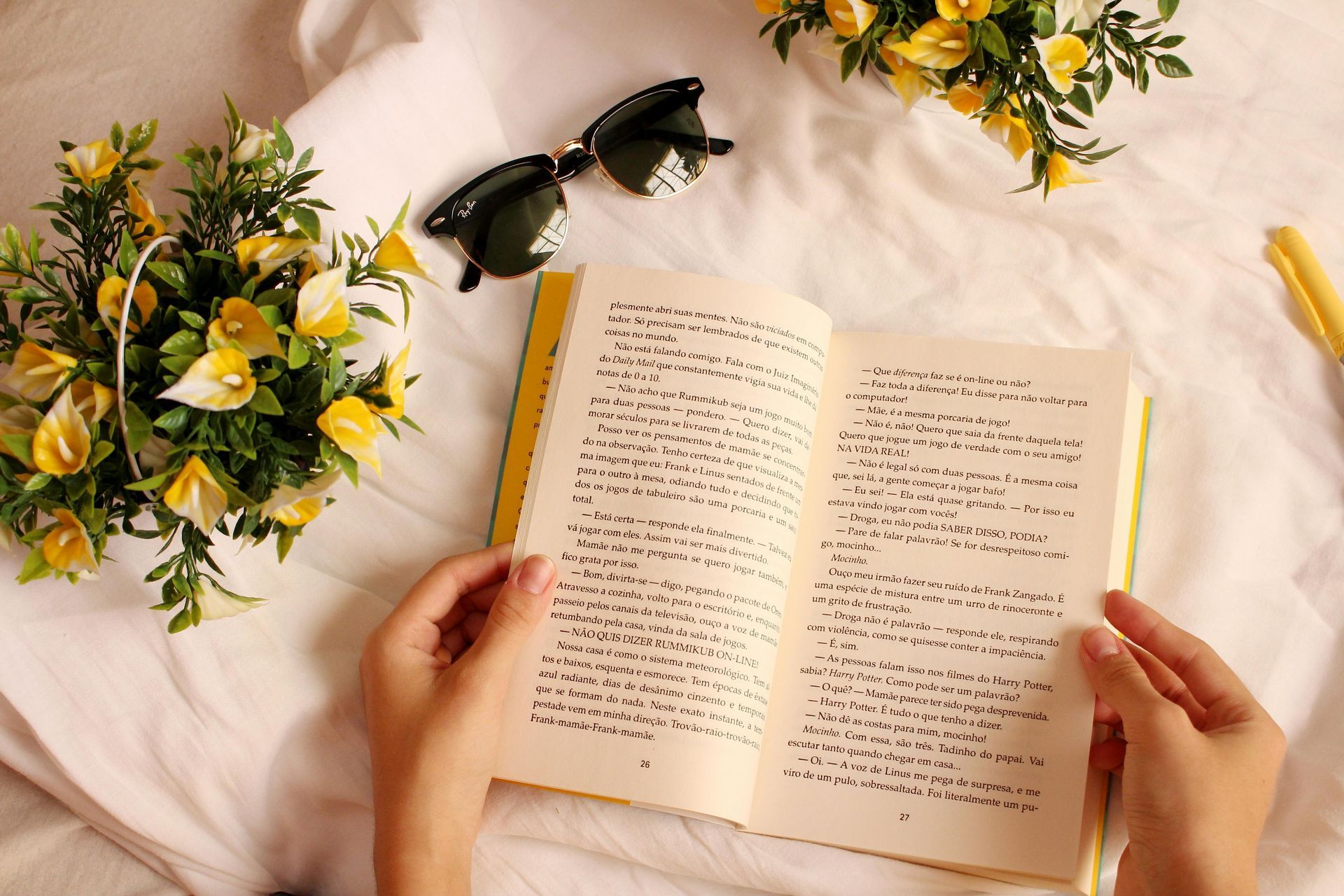 Hands holding open book on white bedding with flowers and sunglasses.