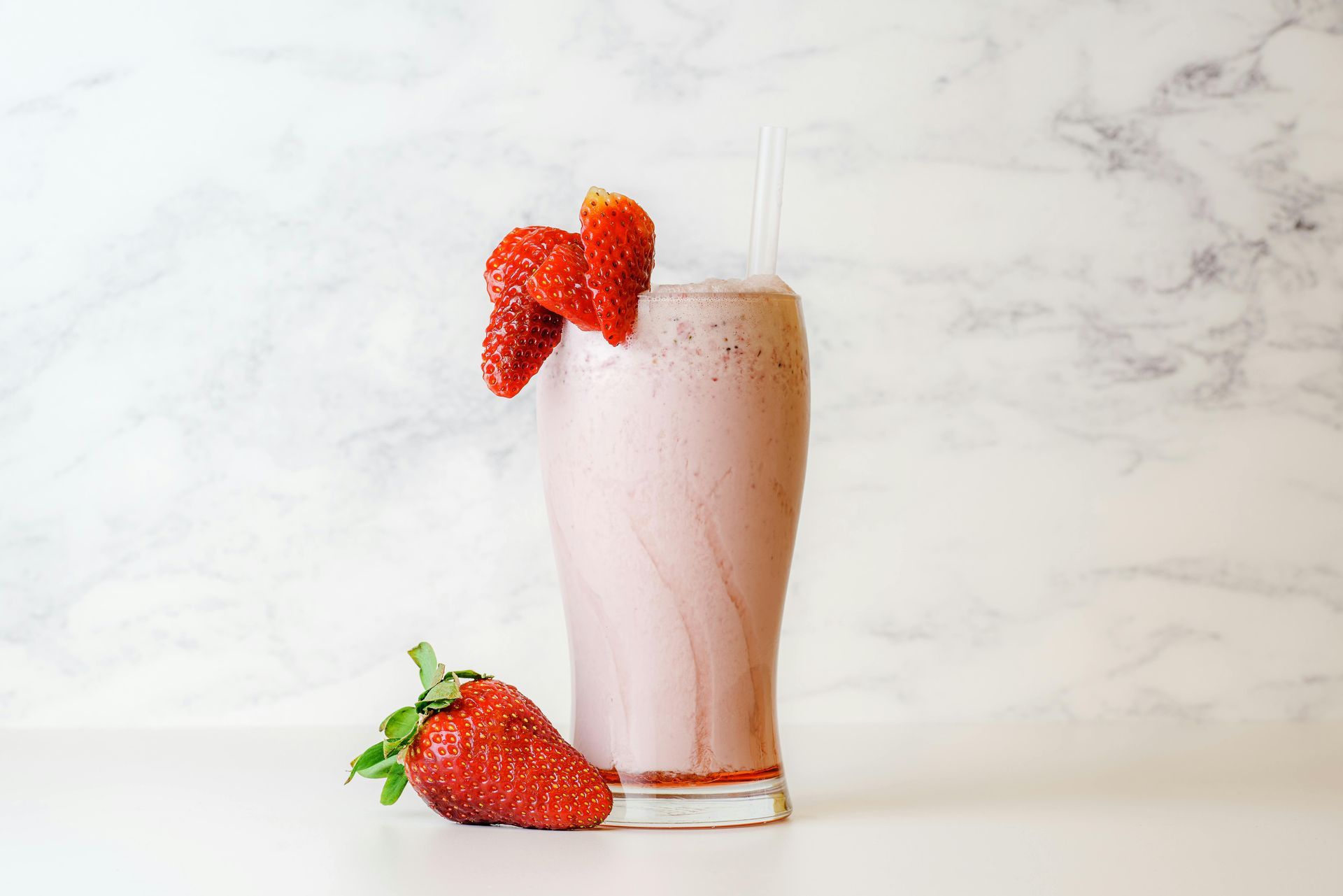 Strawberry milkshake in a glass, garnished with fresh strawberries, on a white surface with a marble background.