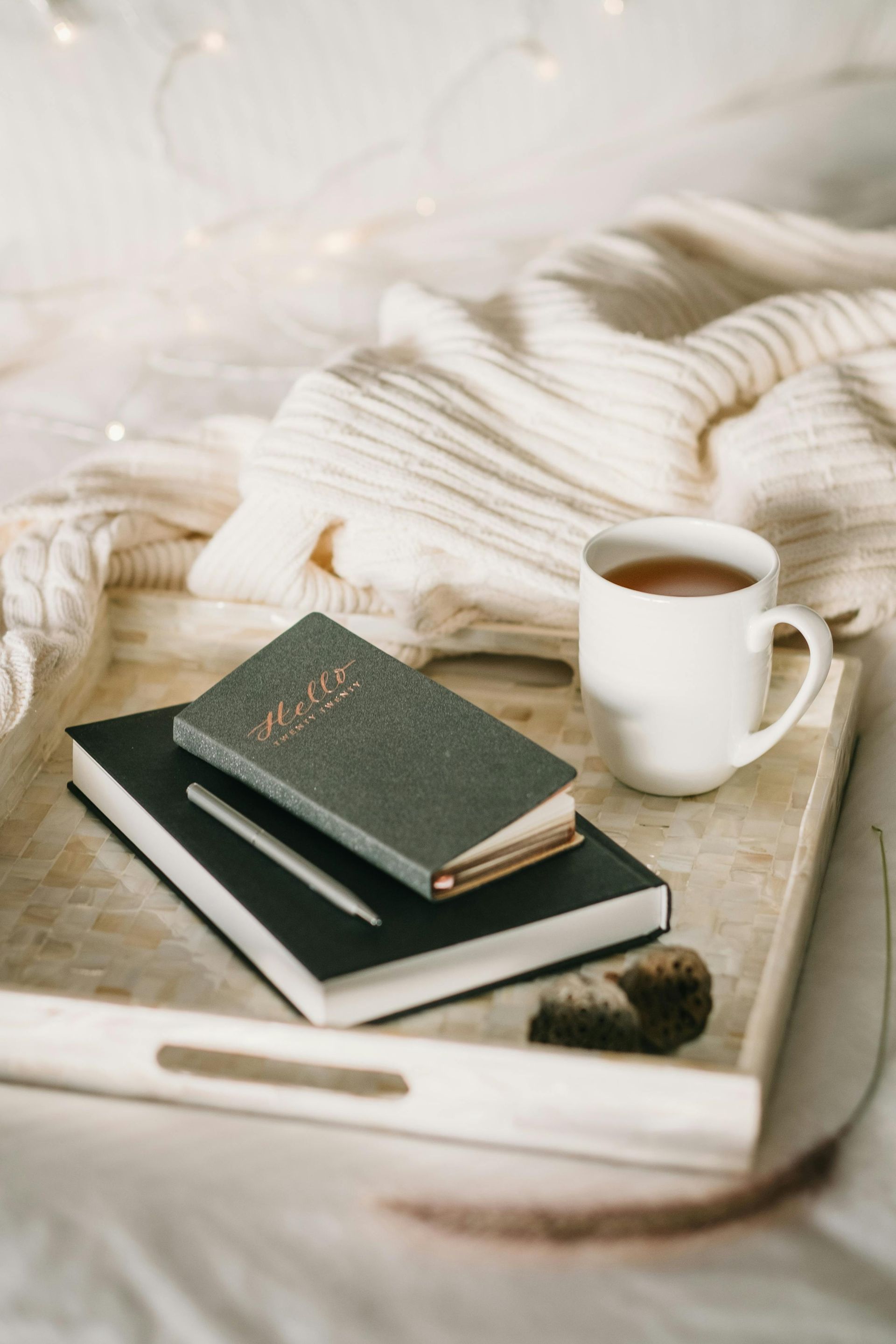 White mug of tea, books, and sweater on a tray on a bed, with soft lighting and a neutral color palette.