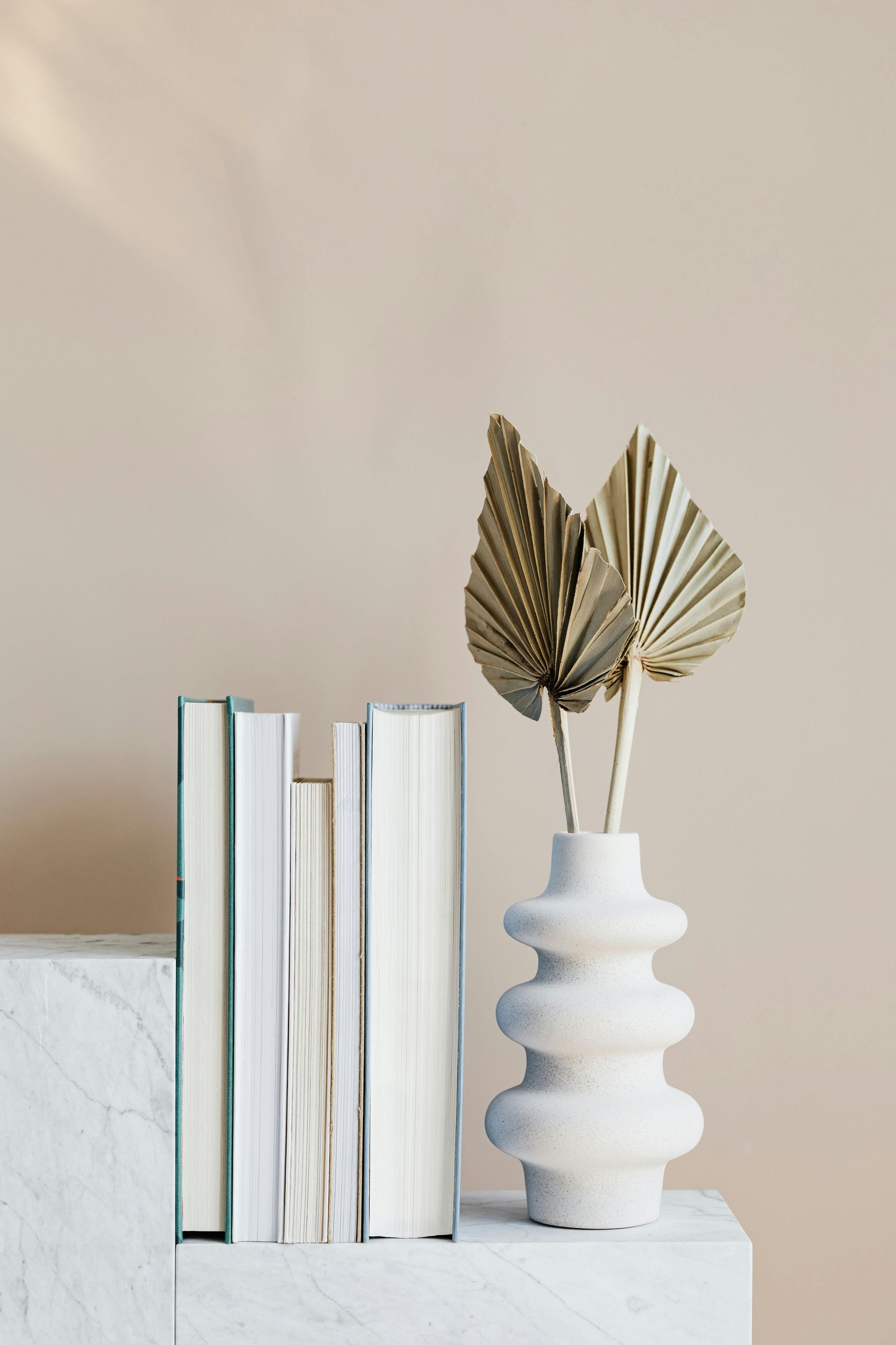 Books and beige vase with dried leaves on a marble block against a neutral wall.