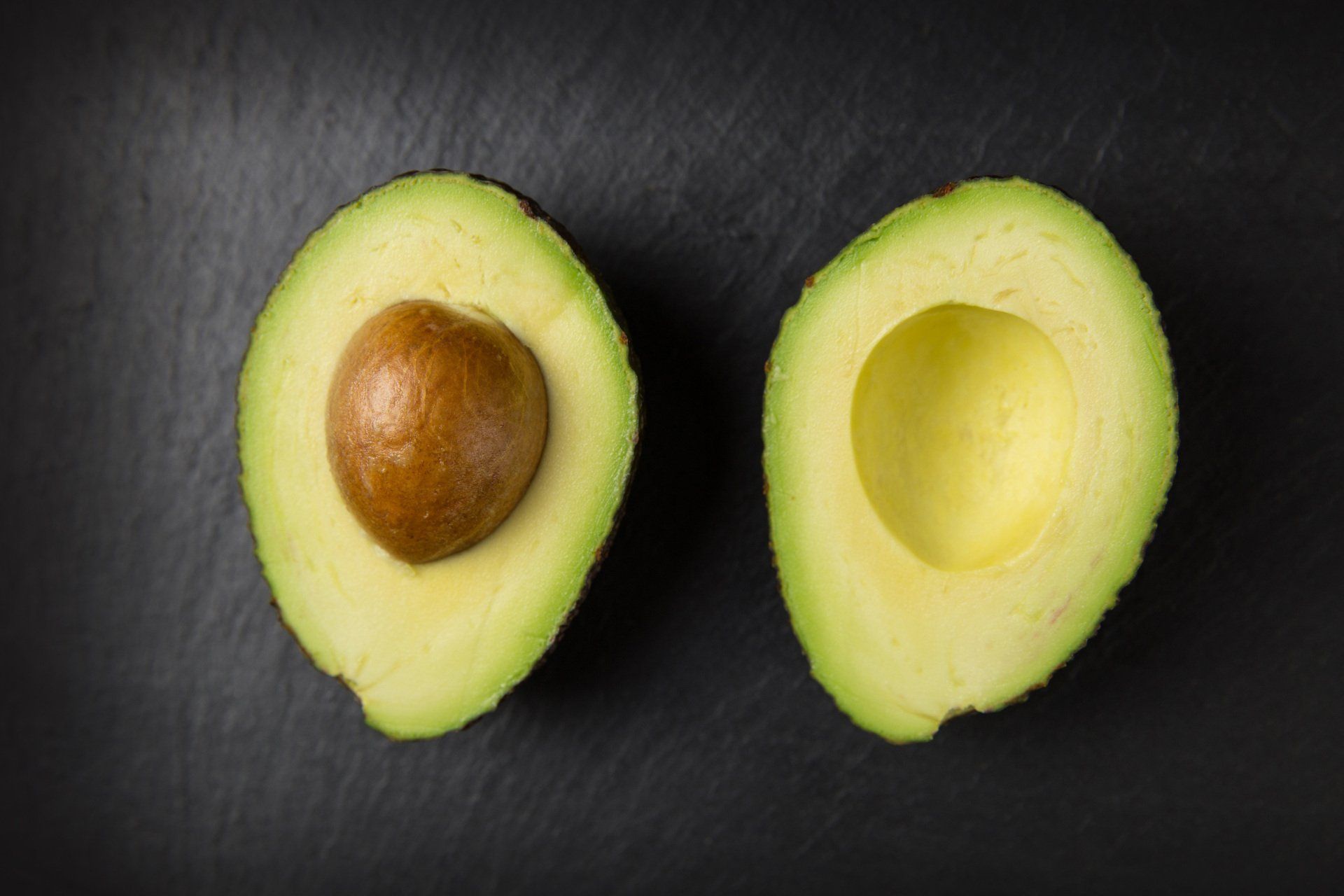 Avocado, cut in half, showing the seed on the left, set on a dark surface.