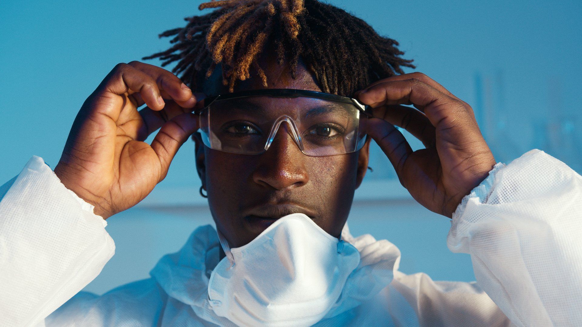Person in lab coat, safety glasses, and face mask, adjusting glasses. Blue background.