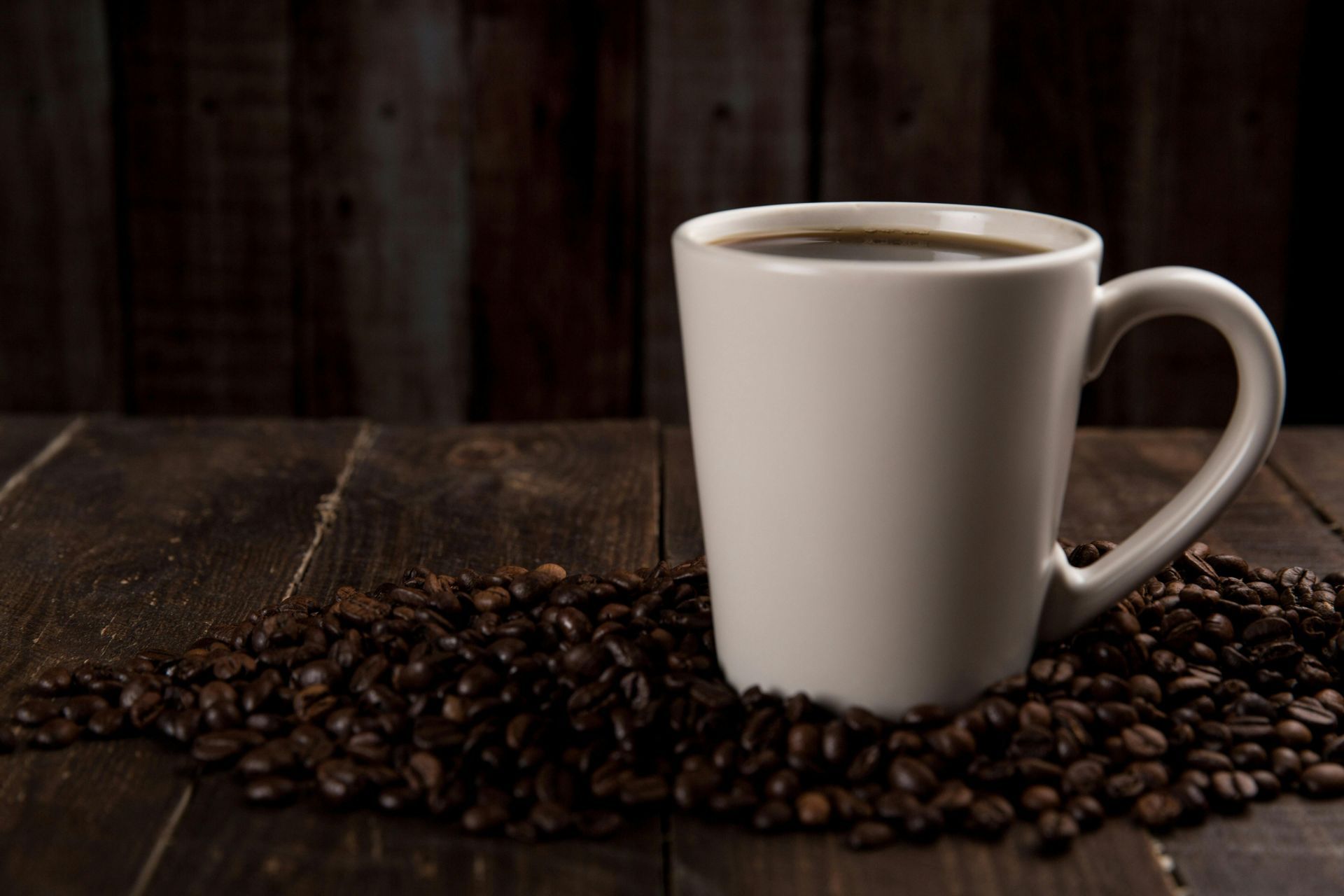 White coffee mug filled with coffee on a bed of coffee beans, against a dark wooden background.