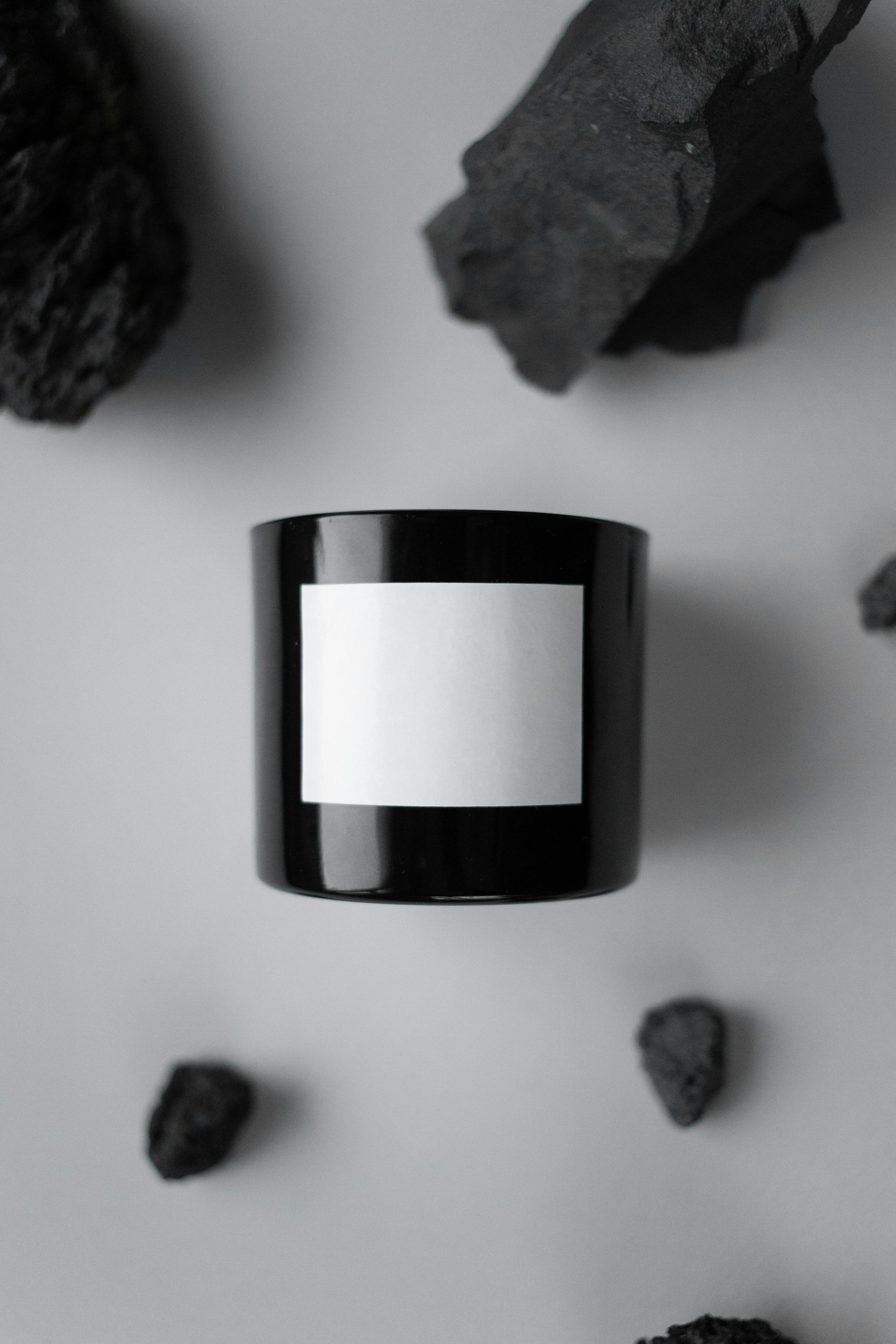 Black candle with a blank white label, surrounded by dark gray rocks on a light gray surface.