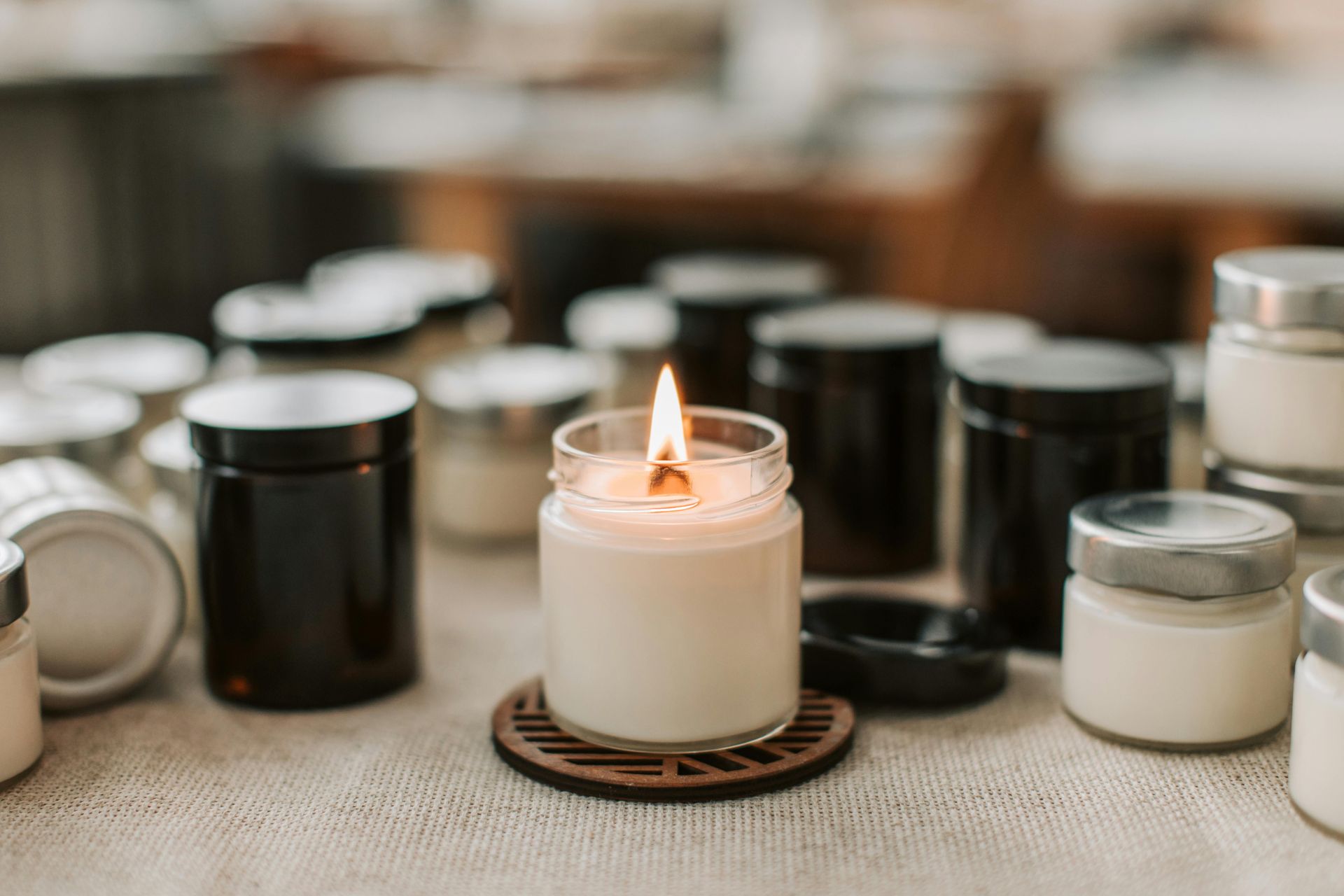 Lit candle on a wooden coaster surrounded by jars of candles on a table.