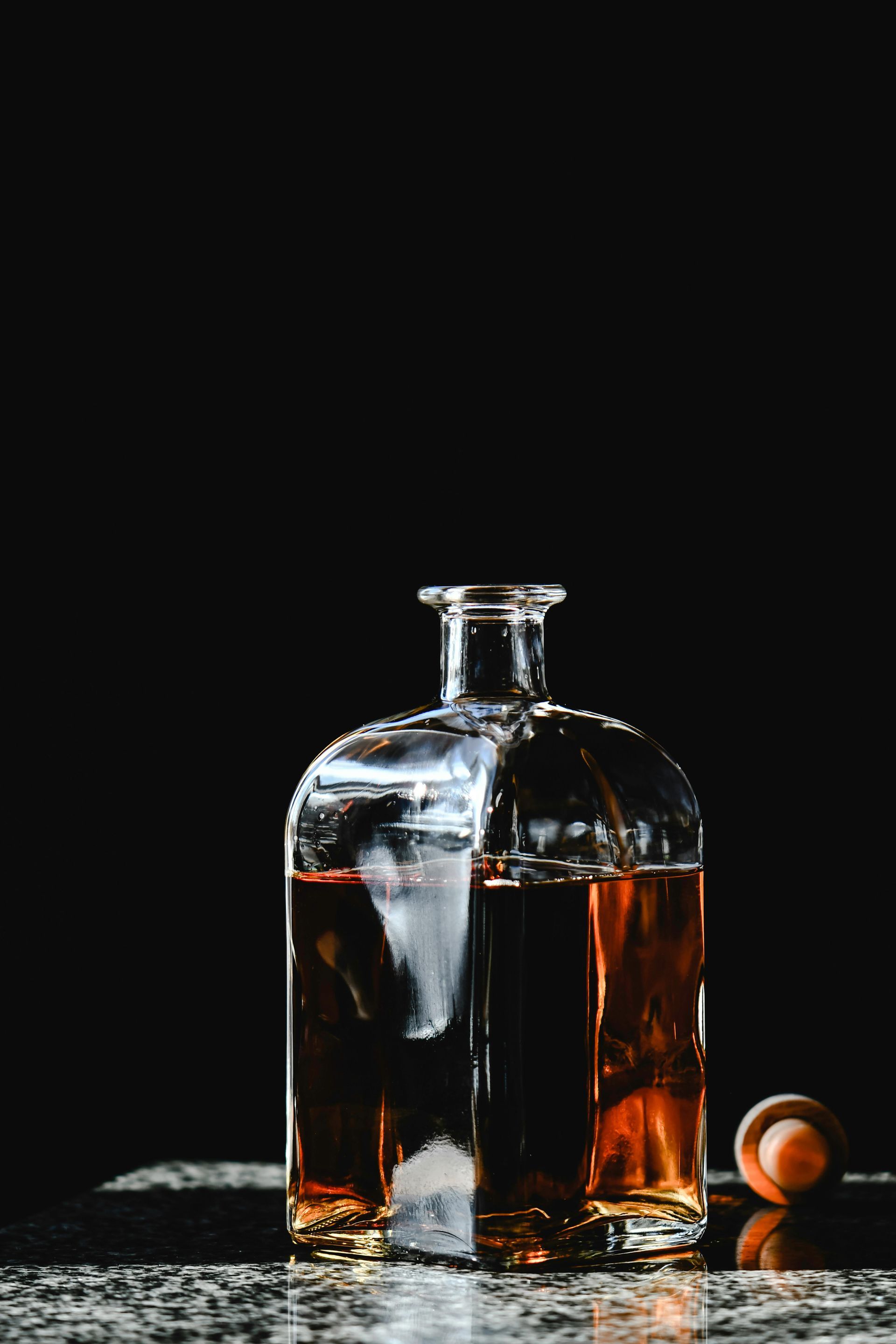 Clear glass bottle with amber liquid, cork beside it, on a reflective surface against a black backdrop.
