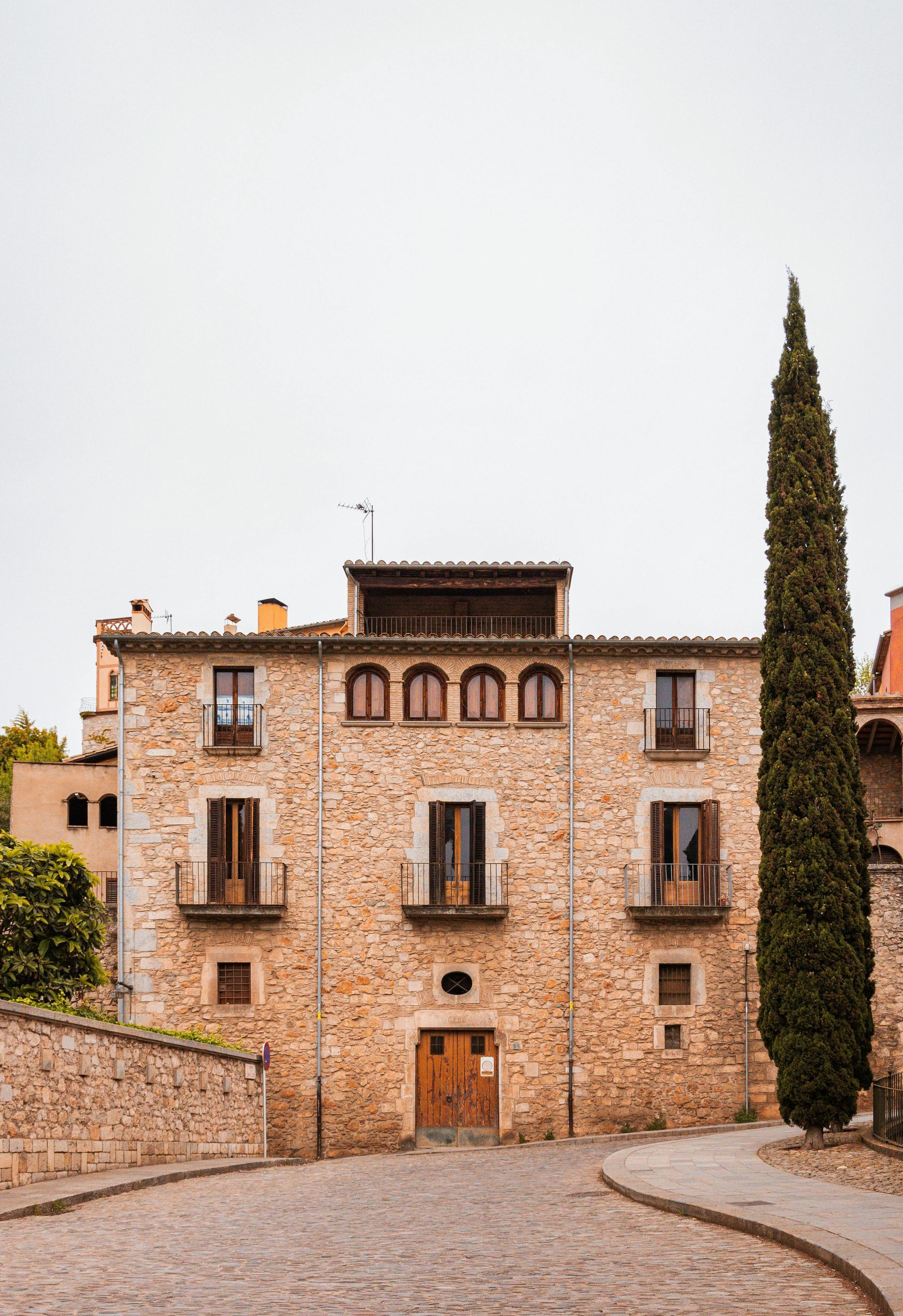 Stone building with arched windows and wooden doors, tall tree alongside a cobblestone street.