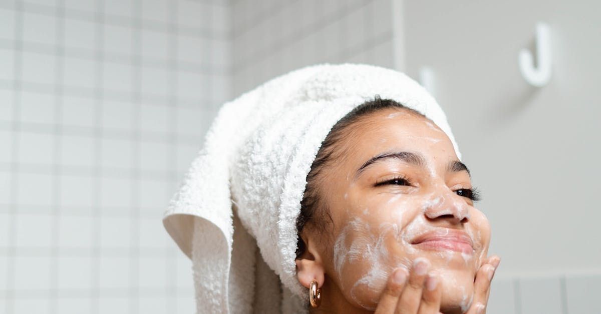 Woman with towel on head, applying facial cleanser in a bathroom.