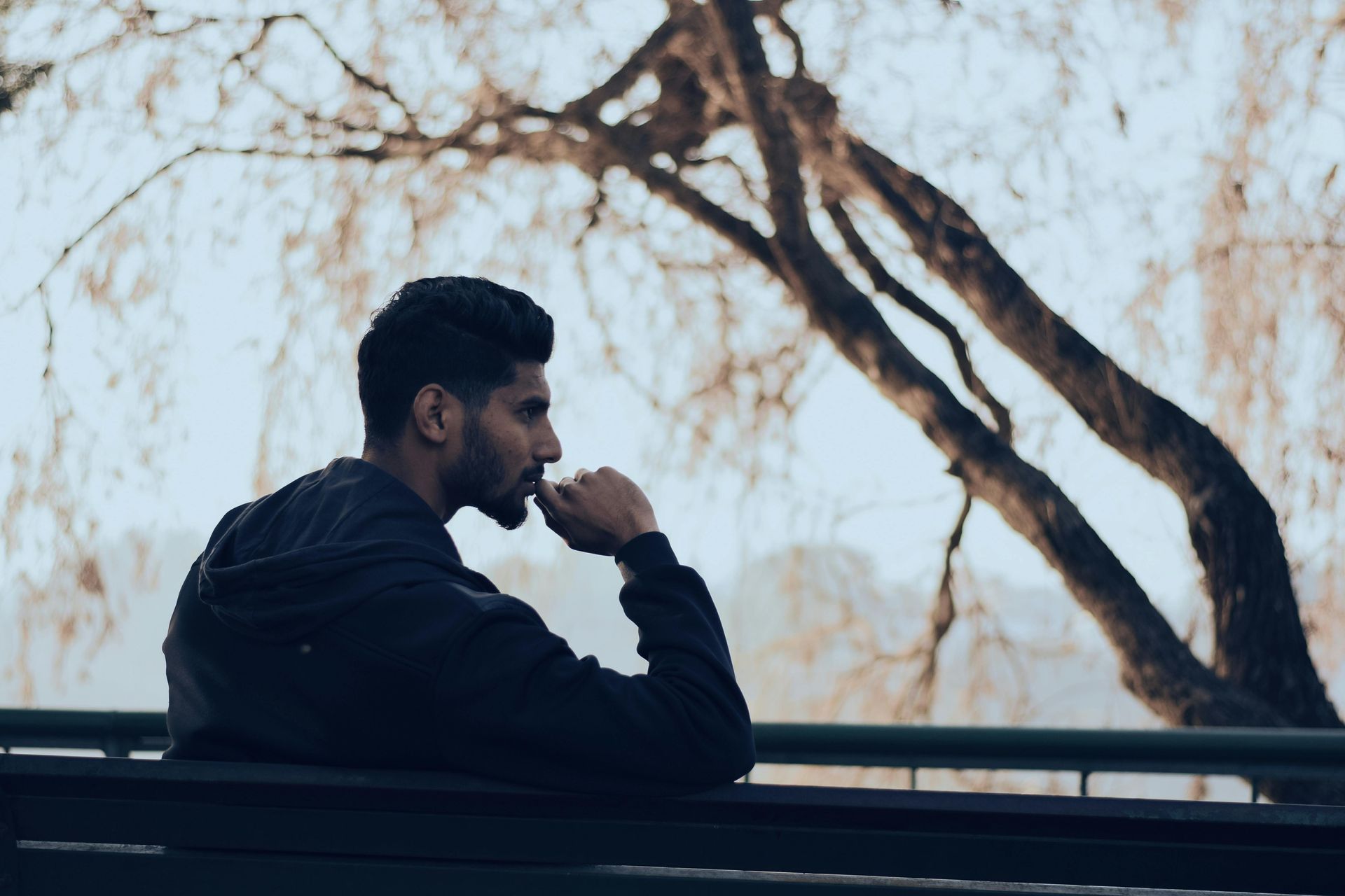 Man in dark jacket sits on a bench, looking thoughtful. Tree branches and overcast sky in background.