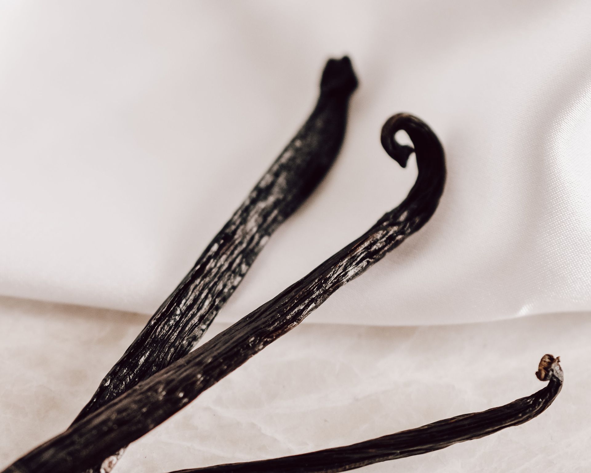 Three dark vanilla beans on a white satin surface.