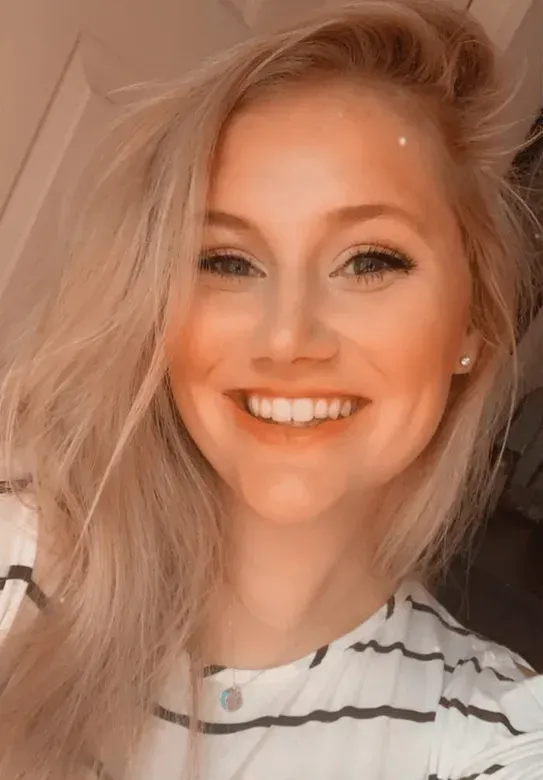 Blonde woman smiles, wearing a striped shirt. Indoor setting with soft lighting.