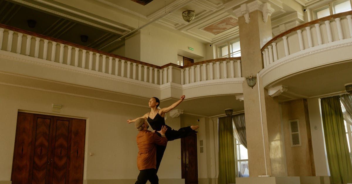 A ballet dancer being lifted in a dance hall with a balcony; the dancer wears a black outfit.