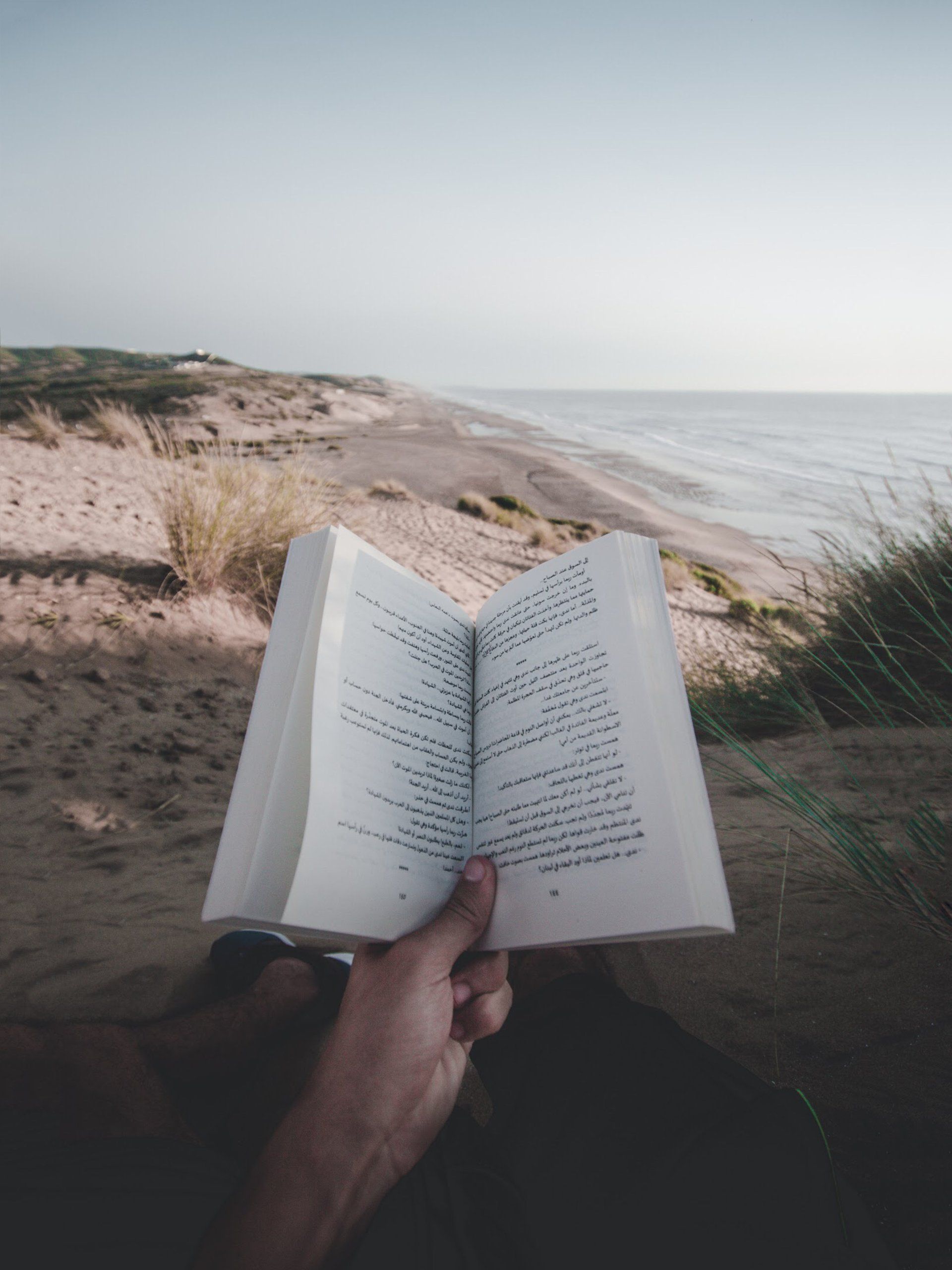 Person holding open book, reading on a sandy beach. Ocean and blue sky in background.