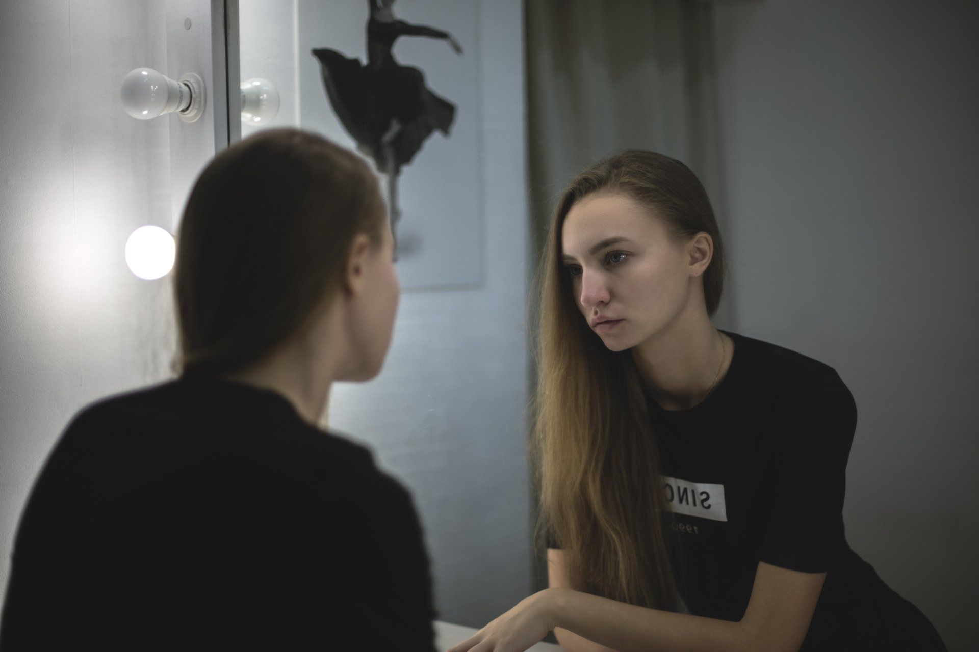 Woman looking at her reflection in a lighted mirror; ballet print on wall.