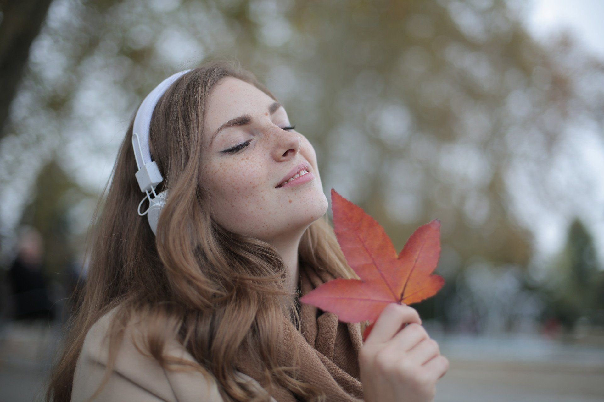 Woman with closed eyes, headphones, and a red leaf, enjoying nature outdoors.