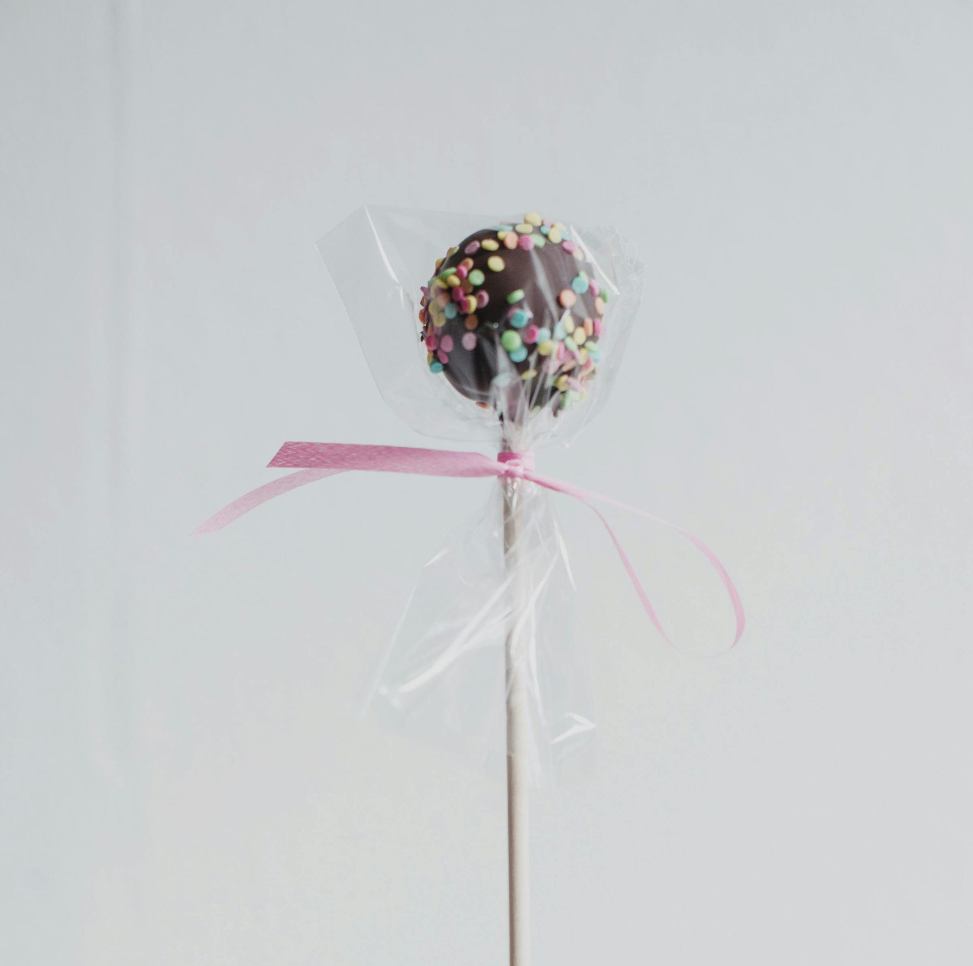 Chocolate cake pop with sprinkles, wrapped in clear cellophane and tied with a pink ribbon.