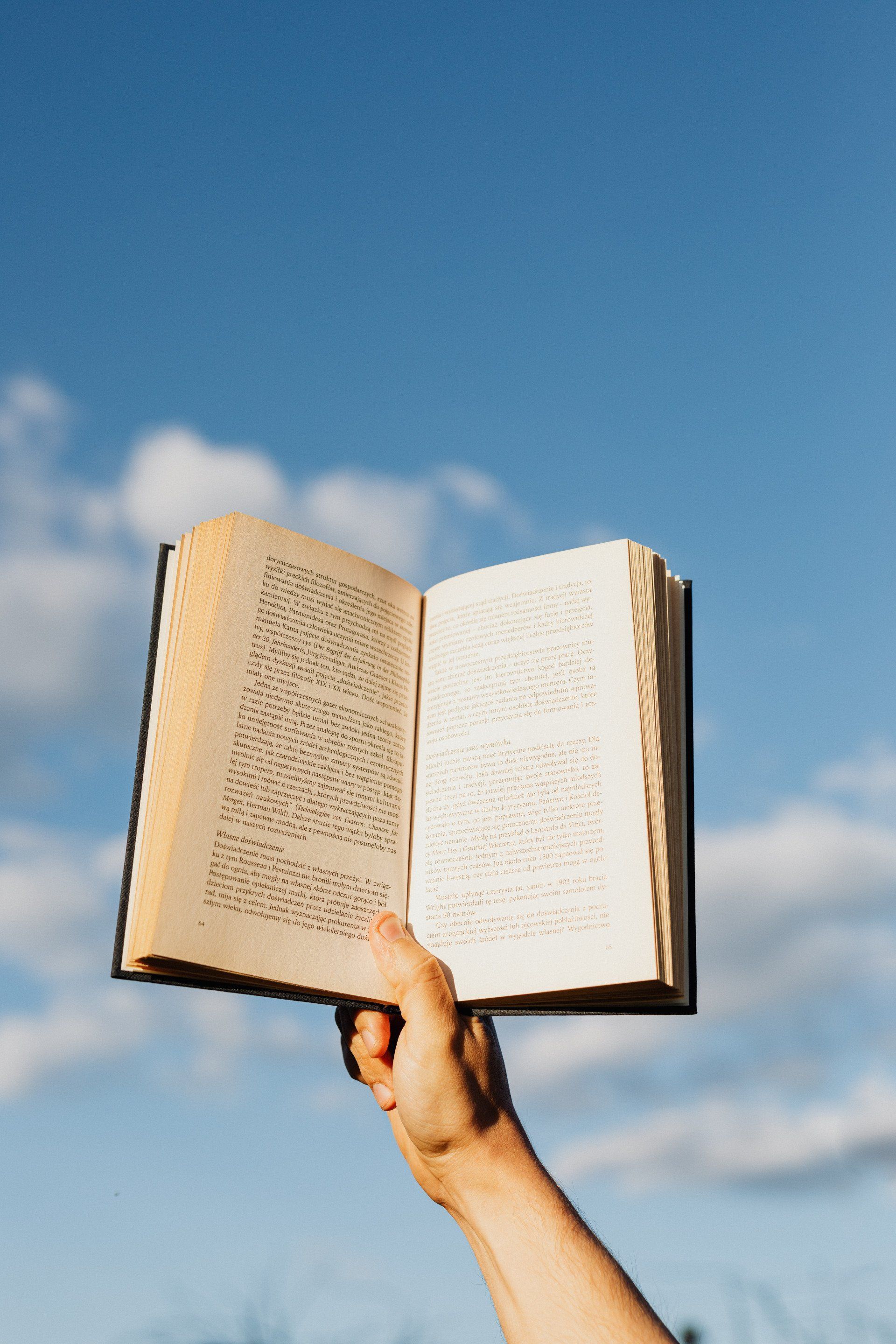 Hand holding an open book against a blue sky with white clouds.