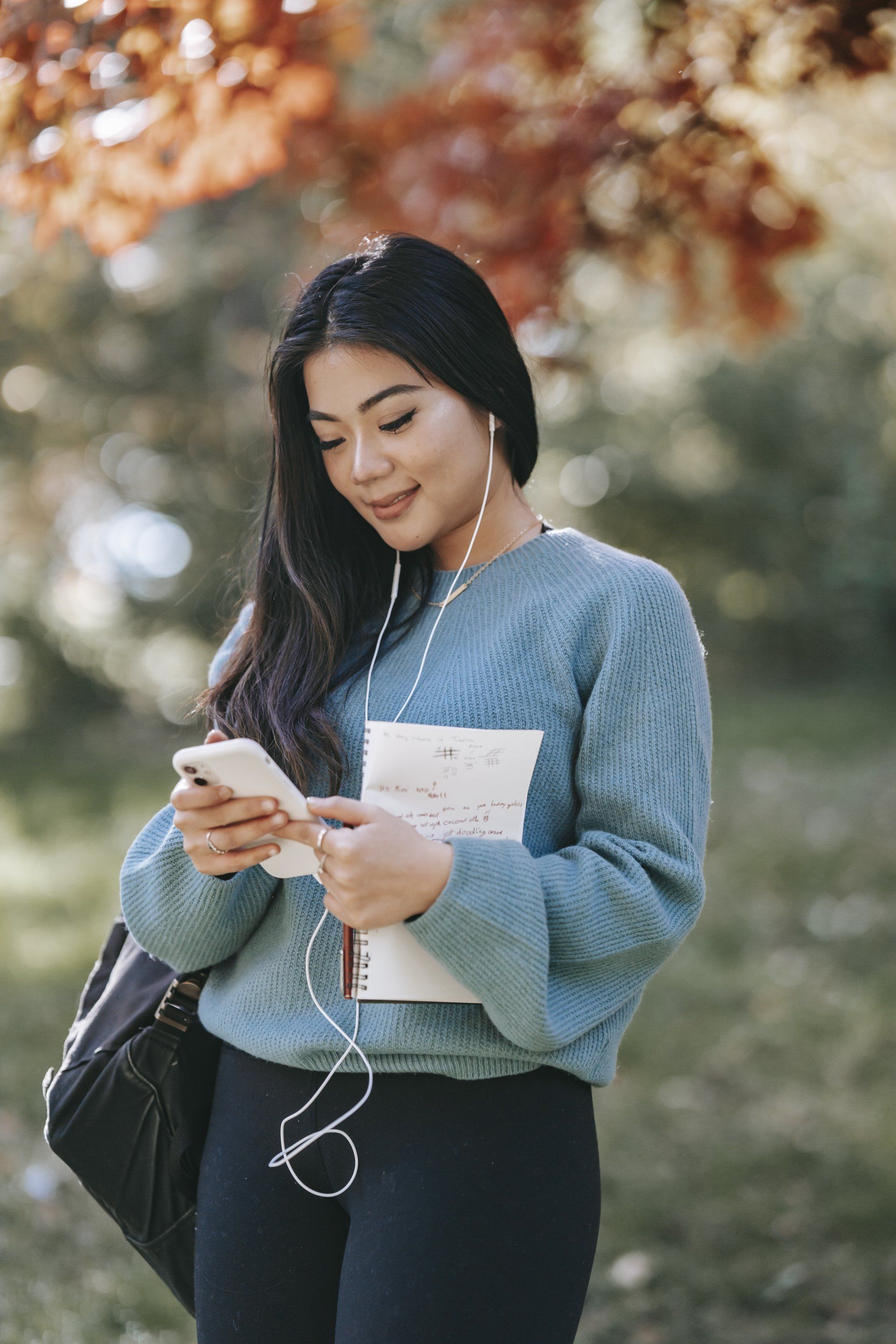 Woman looking at phone, holding notebook, wearing earbuds and a blue sweater, outdoors.