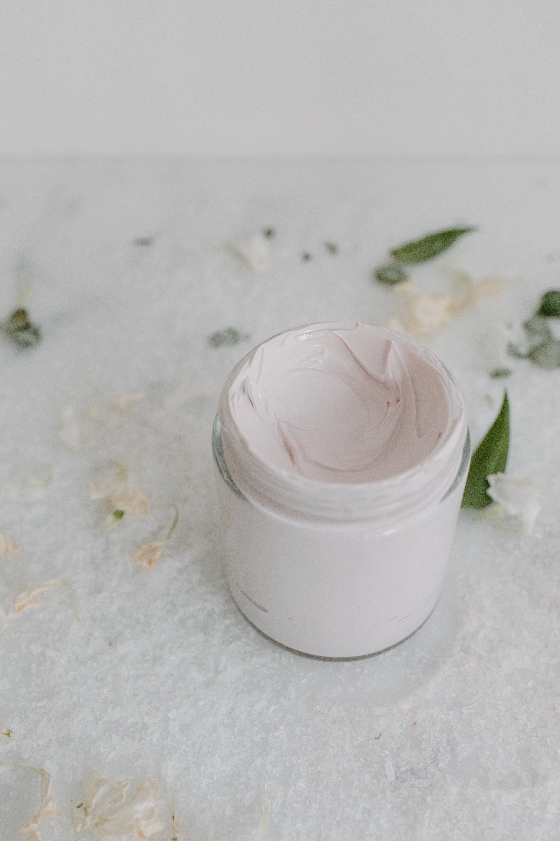 Open jar of pink cream on a light blue surface with scattered flowers and greenery.